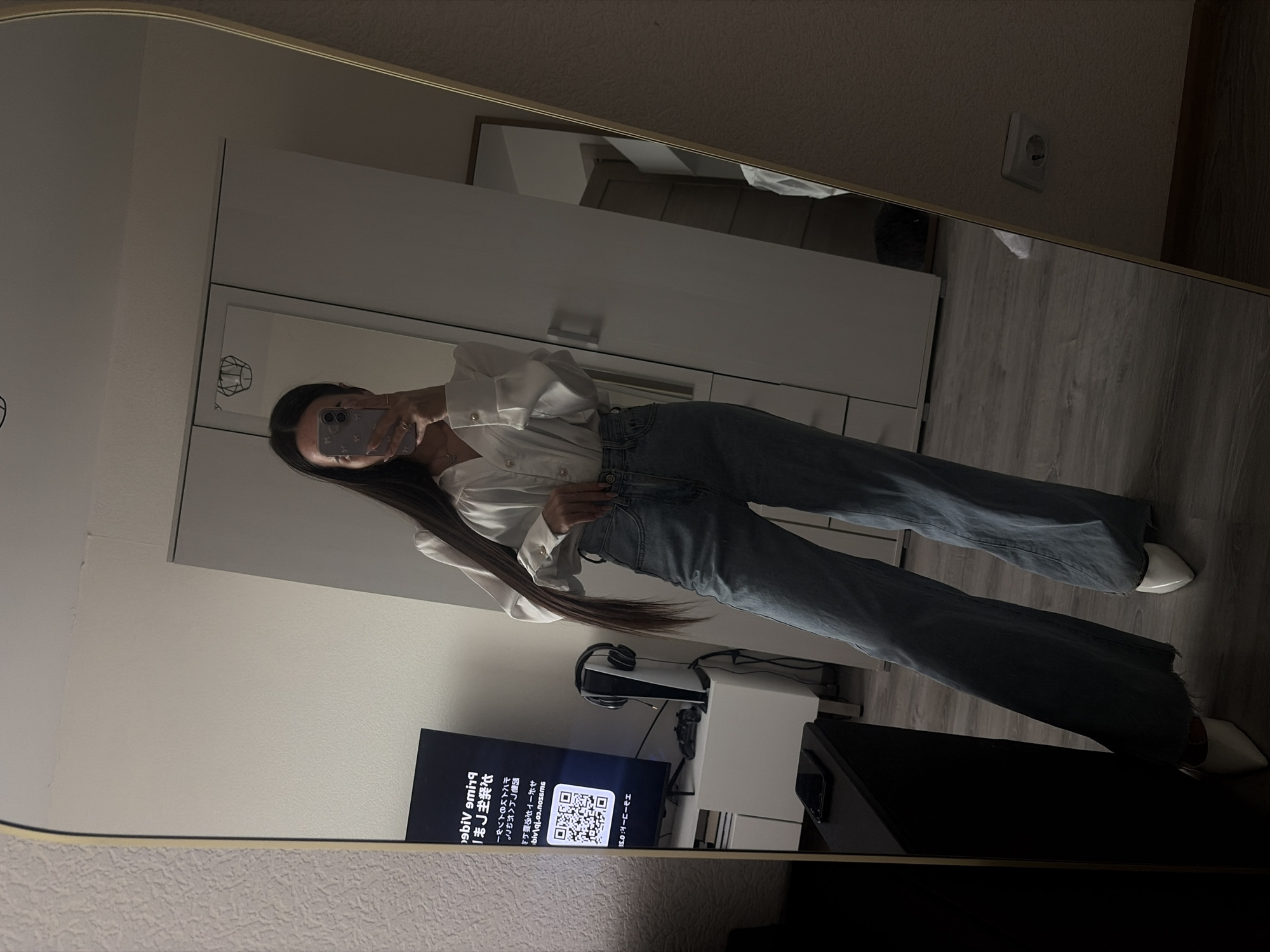 A young woman is captured in a mirror selfie, her image rotated 90 degrees clockwise. She stands before a large, gold-framed mirror, holding a smartphone with a light purple/gray case to take the picture. Her face is partially obscured by the phone, and her long, dark hair cascades over her shoulder. She is dressed in a white button-down shirt, blue jeans, and white pointed-toe heels, presenting her outfit.
The indoor setting, likely a room in Saint Petersburg, Russia, appears modern and furnished. Reflected in the mirror are several items: a white PlayStation 5 console, black over-ear headphones resting on a white stand, and a television screen. The TV displays an Amazon Prime Video activation screen, featuring a prominent QR code and various Japanese characters and text. Visible phrases include "prime vide" (reversed), "J主発," "ヤバトKBノソシー," "つんへがねこ。," and "ヨーローラエ."
Other reflections show a white cabinet or dresser with silver handles, light-colored walls, and wooden flooring. A European-style electrical outlet (Schuko type) is visible on the wall. The ambient indoor lighting is somewhat dim, suggesting the time of day could be evening. The overall scene depicts a casual, personal moment of self-documentation within a contemporary home environment.