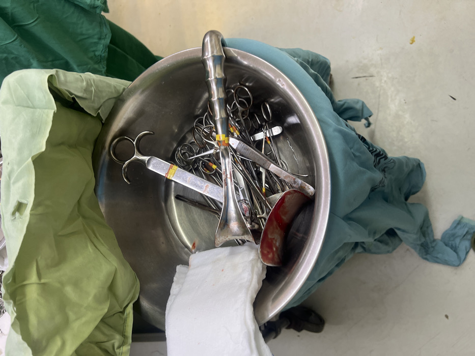 The image captures a close-up, downward-facing view of a stainless steel basin filled with used surgical instruments and soiled textiles, situated within a medical facility in Nairobi, Kenya. The large, round basin contains a dense collection of metal instruments, predominantly forceps and clamps with finger rings, some banded with yellow or orange elastic for identification. A white, folded, cloth-like material, possibly a surgical towel or gauze, is positioned on the left side of the basin, exhibiting a small, distinct red stain. Near the front-left, a curved metal object, appearing to be a retractor blade, is stained with a significant amount of dark red material, consistent with blood. The basin rests upon a draped light blue or teal fabric, which conceals an unseen support structure. Above the basin, to the left, a crumpled light green fabric is visible, possibly a disposable medical drape. The background reveals a light-colored, clean floor surface, and a portion of a dark green fabric in the upper right. A dark shoe is partially visible at the bottom-left, beneath the blue fabric. No individuals are present.
