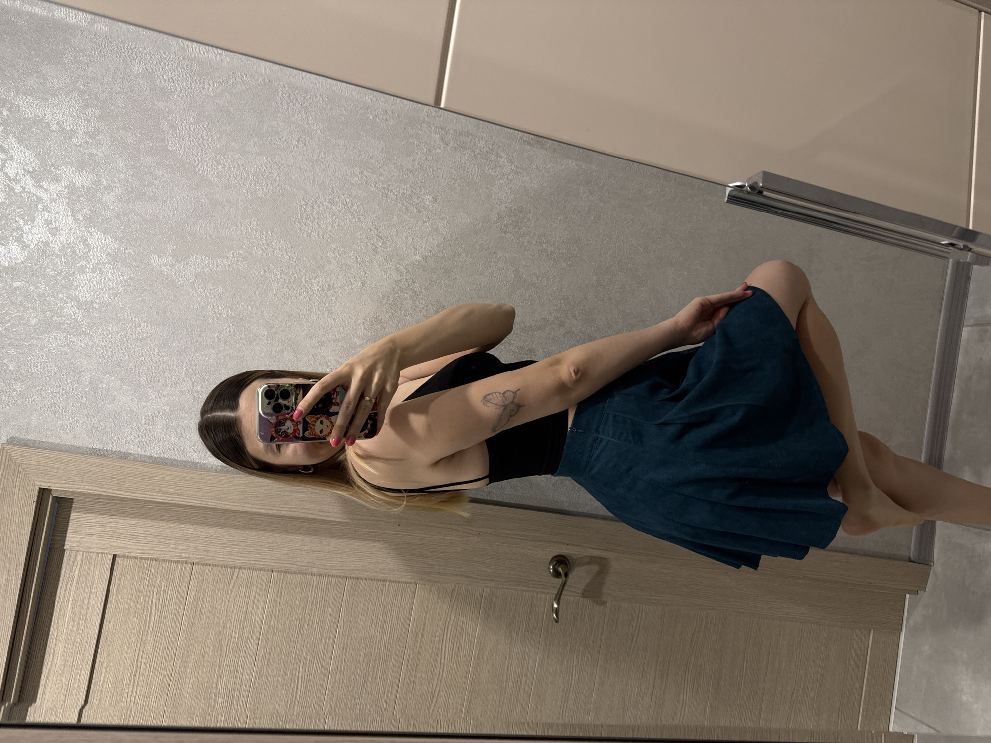 A person is taking a mirror selfie in a room. The person is wearing a black top and a teal skirt. They have long brown hair and a butterfly tattoo on their left arm. The background shows a door and a textured wall. The room appears to be indoors, possibly a bedroom or bathroom. There is no indication of the time of day or weather conditions. The overall mood is casual and relaxed.