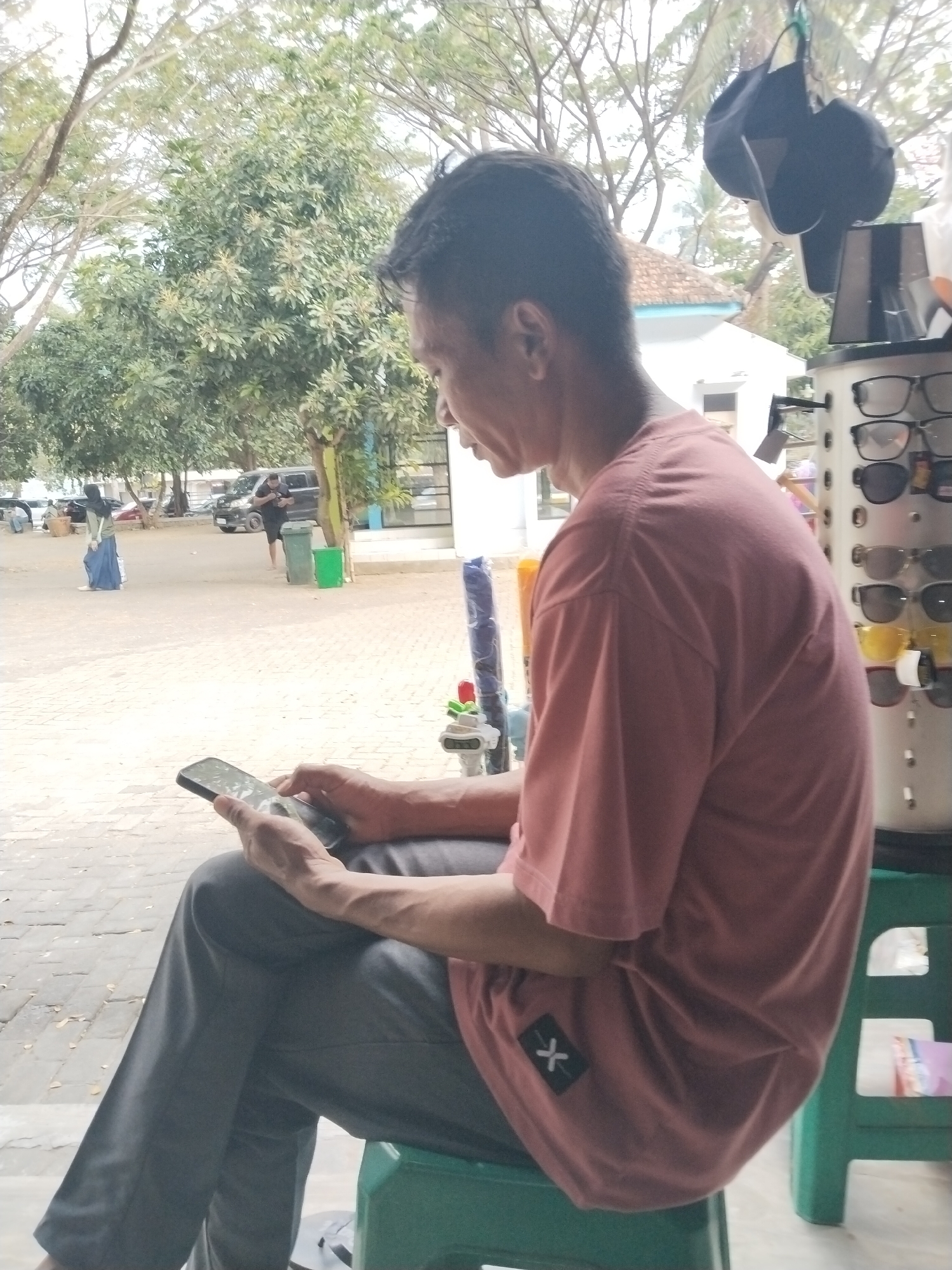 A man is sitting on a green plastic stool, looking at his smartphone. He is wearing a coral-colored t-shirt and grey pants. The man's arms are resting on his lap as he holds the phone. In the background, trees and a white building are visible. A rack of eyeglasses is positioned to the right of the man, with black hats hanging above it. The ground is paved with interlocking bricks, and there are some parked cars and people in the distance, suggesting an outdoor public area. The overall scene appears to be in a commercial area, possibly a shopfront. The location is identified as Prigi, Indonesia.