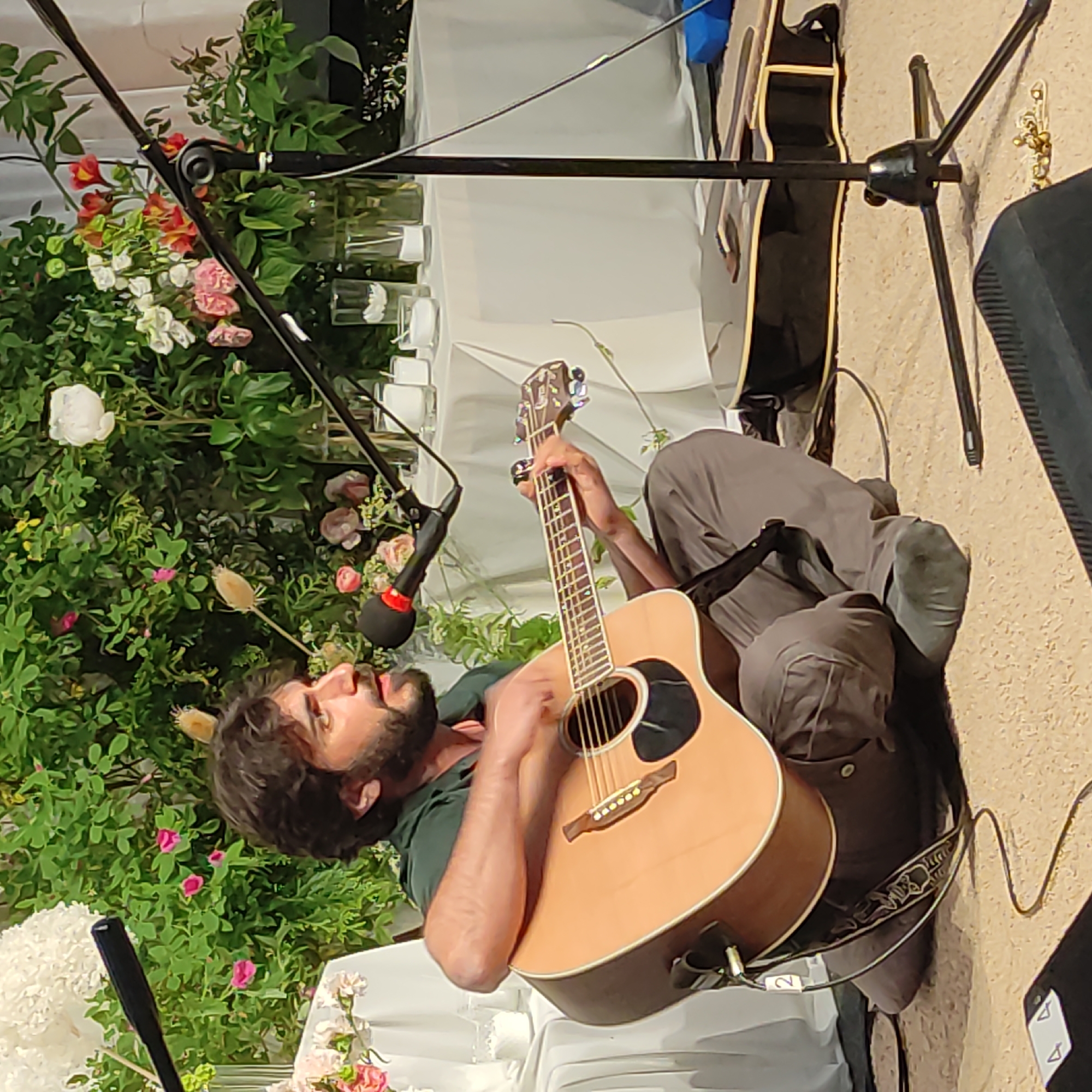 A man with dark, curly hair and a beard is playing an acoustic guitar while sitting on the floor. He is wearing a dark green shirt and grey pants. A microphone is positioned in front of him, and he appears to be singing or speaking into it. The background consists of a white tablecloth draped over a table, adorned with a bouquet of white and pink flowers and some greenery. Several glass vases with flowers and some candles are also visible on the table. The setting appears to be an indoor event, possibly a performance or a gathering. The image is a medium shot, focusing on the man and his guitar, with the background slightly blurred. The lighting is soft and warm. There are no discernible elements that would indicate a specific location in Zhirnosovo, Russia.