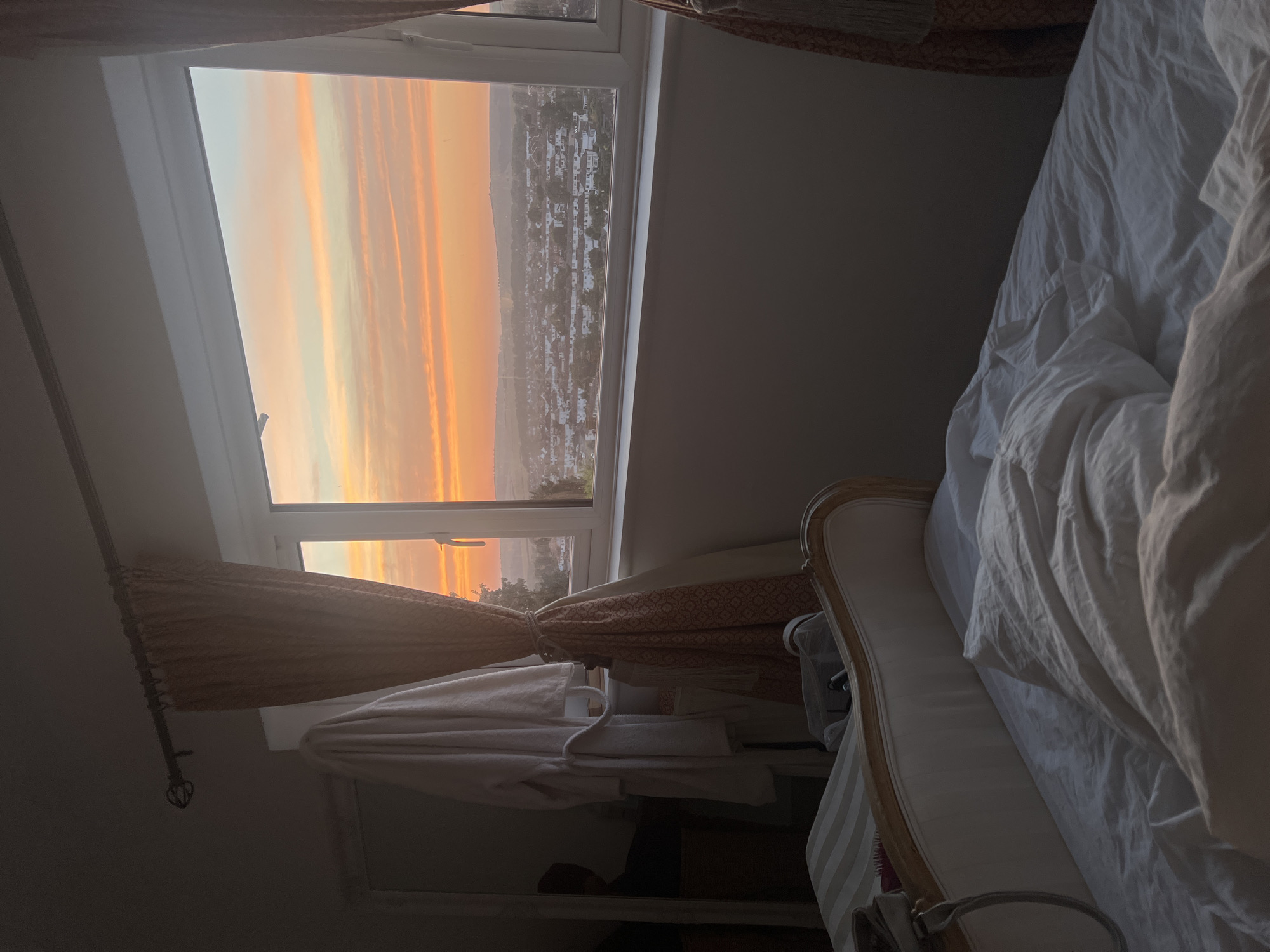 The image depicts a bedroom scene in Brighton, United Kingdom, with a focus on the view from a window at sunrise. The bed is unmade with rumpled white sheets and a duvet, suggesting it has been recently occupied. To the left of the bed, a white bathrobe hangs on a hook, and a floor-length mirror with a carved wooden frame is visible. A patterned curtain, tied back with a rope, partially obscures the window.
The window reveals a panoramic view of a residential area in Brighton as the sun rises. The sky is awash with vibrant hues of orange, pink, and yellow, with streaks of pastel blue. The city below is dotted with houses and buildings, nestled on rolling hills. The light suggests an early morning in a temperate climate. The overall atmosphere is serene and tranquil, capturing a quiet moment before the city fully awakens.