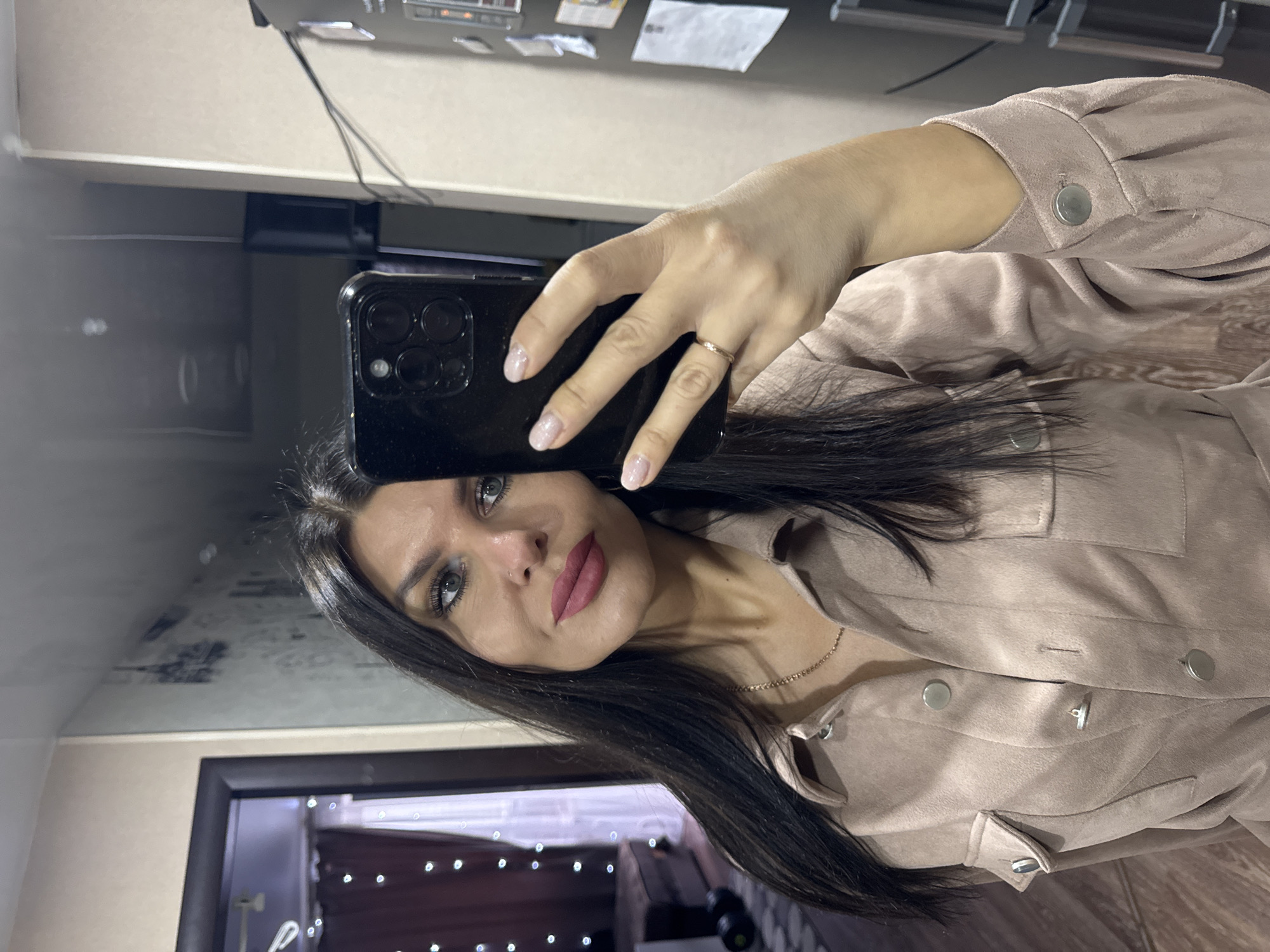 A woman takes a mirror selfie indoors, holding a black smartphone with her right hand. The image is oriented with her head to the left and body extending to the right. She has long, dark hair and light-colored eyes. Her face is made up with dark eyeliner/mascara and dark pink lipstick, and she has a small nose piercing. She wears a simple gold-colored ring on her right ring finger and a thin gold-colored chain necklace. Her nails are manicured with a light, possibly glittery, polish.
She is wearing a beige or light brown long-sleeved shirt or jacket, possibly made of a suede-like material, featuring silver snap buttons and visible seam details. The sleeves appear slightly puffed at the shoulders.
The background is a reflection of the room she is in. Above her head, a light-colored wall is visible, with a darker shelf or counter just above her phone. Further back, in the upper-left of the reflection, there are some papers or notes affixed to a surface, though any text is illegible. The lower part of the background shows a dark framed opening, possibly a doorway or window, with blurred lights resembling string lights or indoor decorations, suggesting an evening or nighttime setting. The overall ambiance is indoor. No specific location cues for Chaikovsky, Russia are discernable from the image.