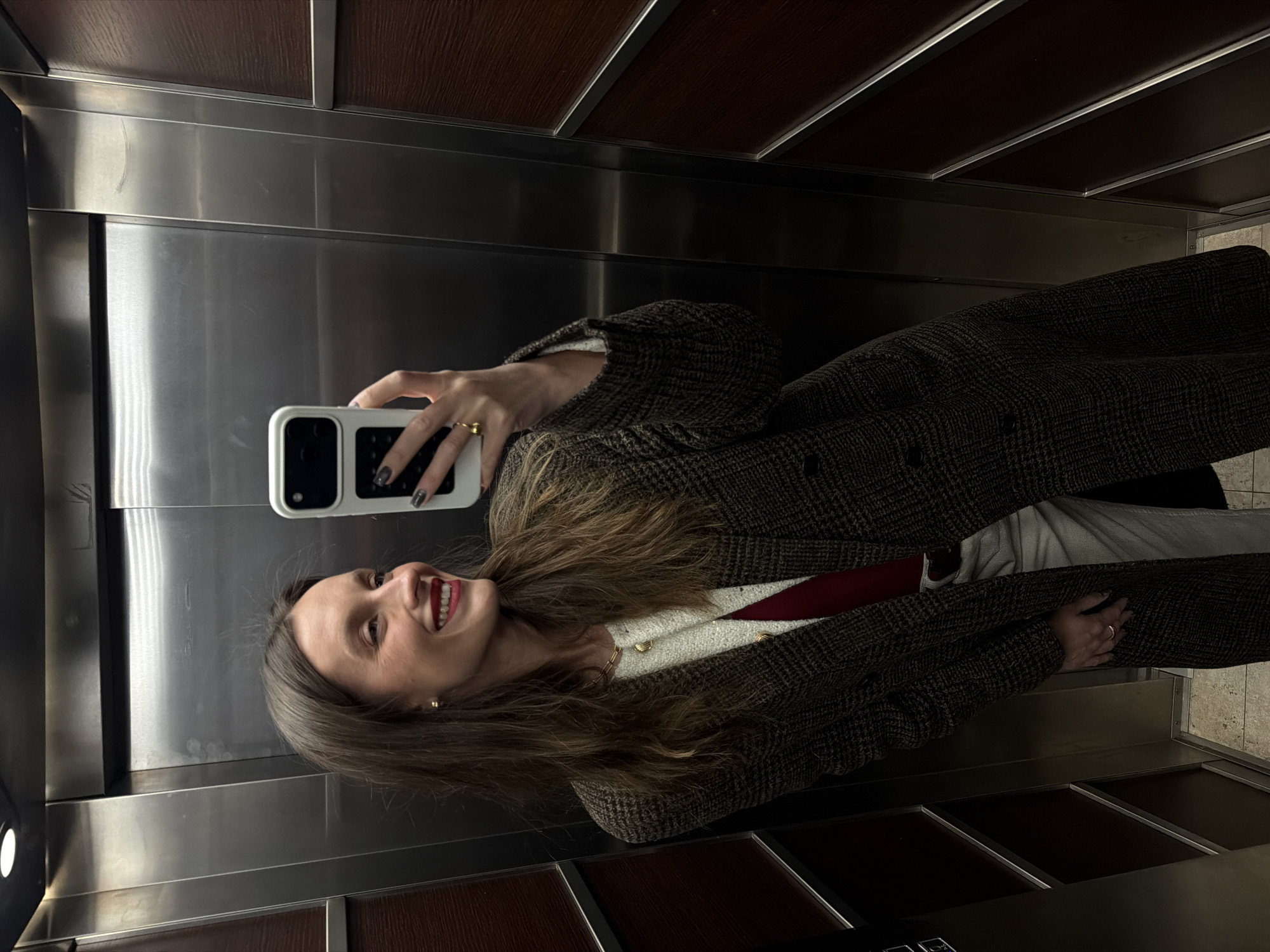 A woman is taking a selfie in what appears to be an elevator. She is wearing a brown plaid coat over a light-colored top with a visible red element, possibly a scarf or sweater. She has long brown hair, fair skin, and is smiling. Her left hand is holding a smartphone with a white case and the fingers of her right hand are visible. The elevator's interior consists of brushed metal walls with dark brown wood paneling at the top and bottom. The location is not discernible from the image.