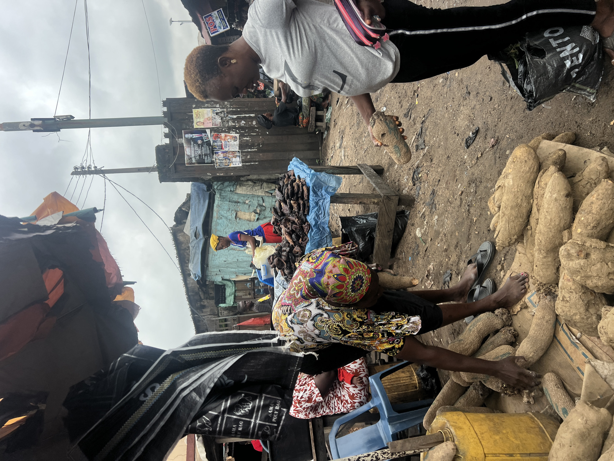 In Nkpor, Nigeria, a street market bustles with activity. Two women are the primary subjects. One woman, wearing a grey t-shirt and black leggings, holds up a large yam for inspection. She has blonde hair and gold earrings. The other woman, wearing a colorful headwrap and a patterned shirt, is bent over a pile of yams, appearing to select or arrange them. She wears black shorts and flip-flops. In the background, a person in a yellow headwrap and blue shirt is visible behind a table laden with dried fish. The market is characterized by makeshift stalls with tarpaulin roofing. Piles of yams are a prominent feature of the scene, both on the ground and on a wooden table. Various objects, including plastic bags, a yellow container, and chairs, are scattered around the market. The ground is dirt and appears somewhat muddy. Electrical wires and poles are visible overhead, suggesting an urban setting. Posters are plastered on a corrugated metal wall in the background. The sky is overcast.