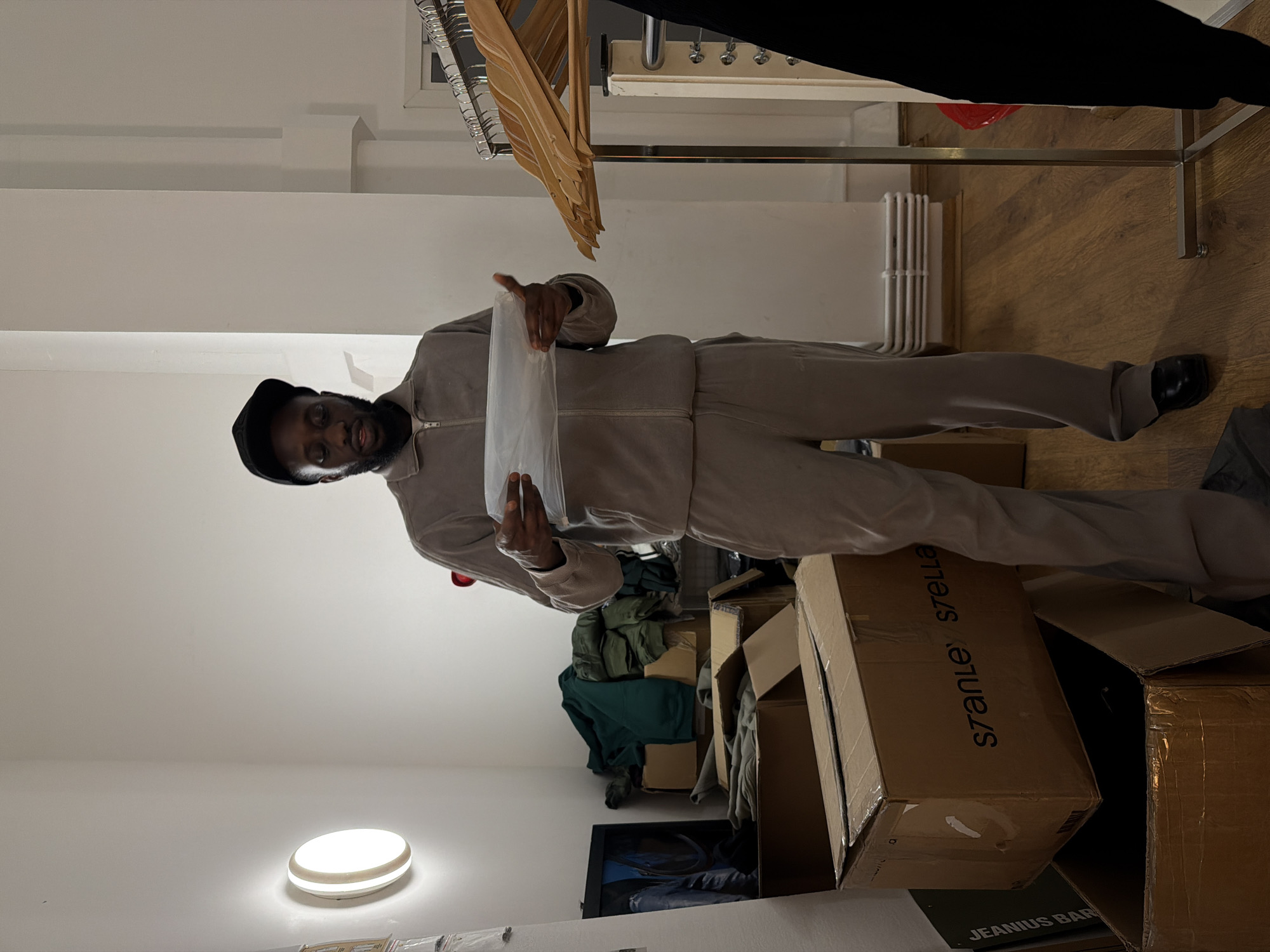 The image captures an indoor scene, likely a stockroom, back office, or a retail space undergoing setup. A man stands centrally in the frame, directly facing the camera with a pleasant, slight smile. He has dark skin, a beard, and is wearing a dark cap and a light grey or beige full-zip jumpsuit or tracksuit. His left hand, adorned with a ring on the ring finger, and his right hand are holding up a clear plastic bag or document.
The room features white walls and a wooden floor. In the background, to the man's left, metal clothing racks with wooden hangers are visible, some holding dark garments. A white radiator is mounted on the wall further back. The lower portion of the image shows several cardboard boxes on the floor. One prominent brown box features the text "s7anley stella" (likely "Stanley/Stella"). Another box, partially visible in the bottom right corner, has "JEANIUS BAR" printed on it. Piles of clothing, including green and darker items, are visible among the boxes and on the floor. A round, white ceiling light fixture with a warm glow illuminates the space from the lower left side of the frame. The overall impression is that of a functional working environment where items are being organized or prepared.