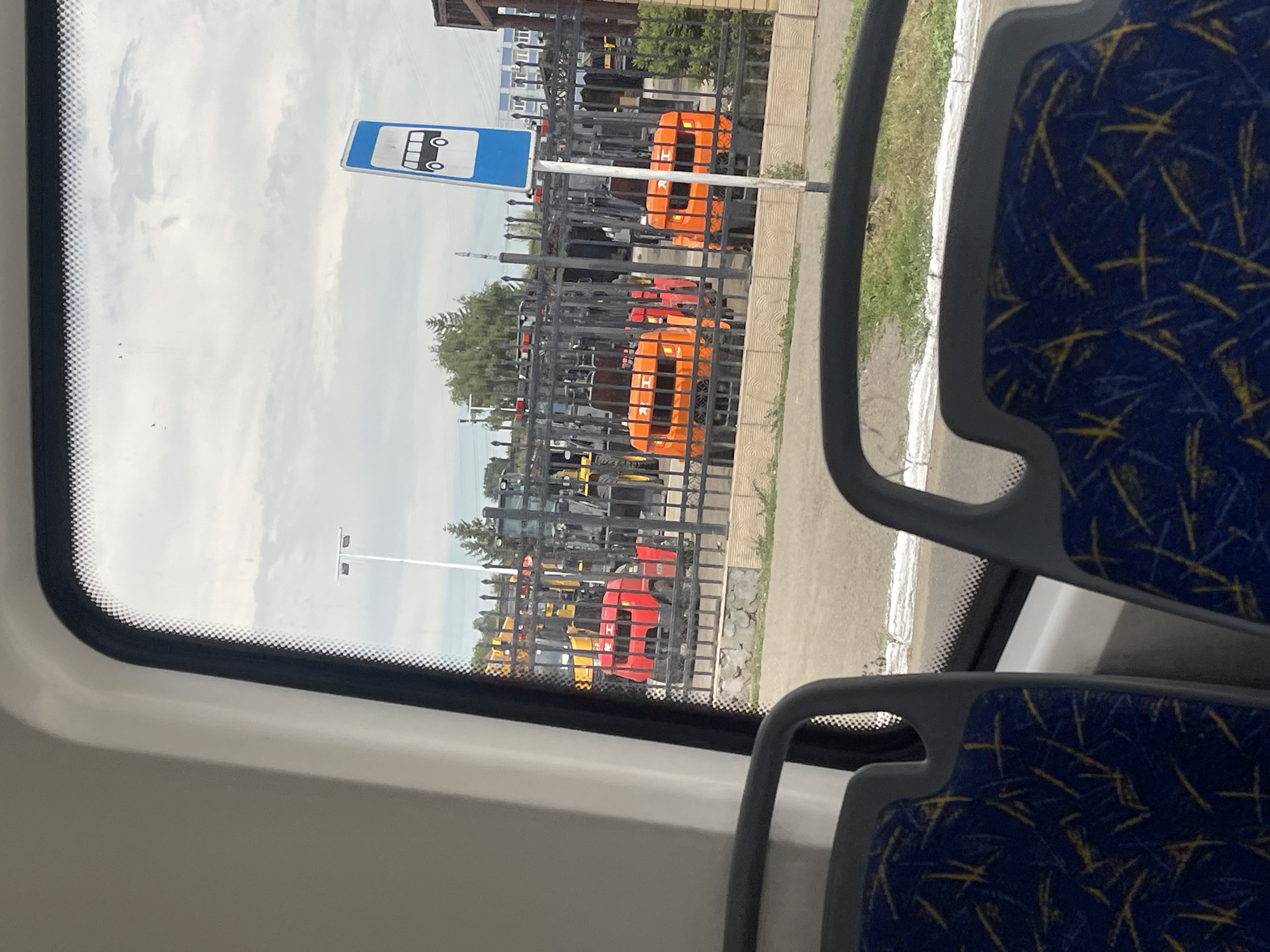 The image, captured from inside a vehicle, offers a view through a window onto an outdoor scene. In the foreground, the patterned blue and yellow upholstery of seats is visible, suggesting the interior of a bus or similar public transport. Beyond the seats, the window frames the exterior.
The exterior scene depicts a fenced area containing several industrial vehicles, primarily forklifts, in various colors including red and orange, with some yellow machinery also present. The forklifts appear to be parked or stored. Behind the fence, a bus stop sign with a blue background and a white bus icon is prominently displayed, indicating this location is likely a public transportation stop. Further in the background, trees and buildings, possibly industrial in nature, are visible under a cloudy sky. The presence of industrial equipment and the bus stop sign suggest an area that serves both industrial and public transport needs. The context provided indicates the image was taken in the city of Oskemen, Kazakhstan.
