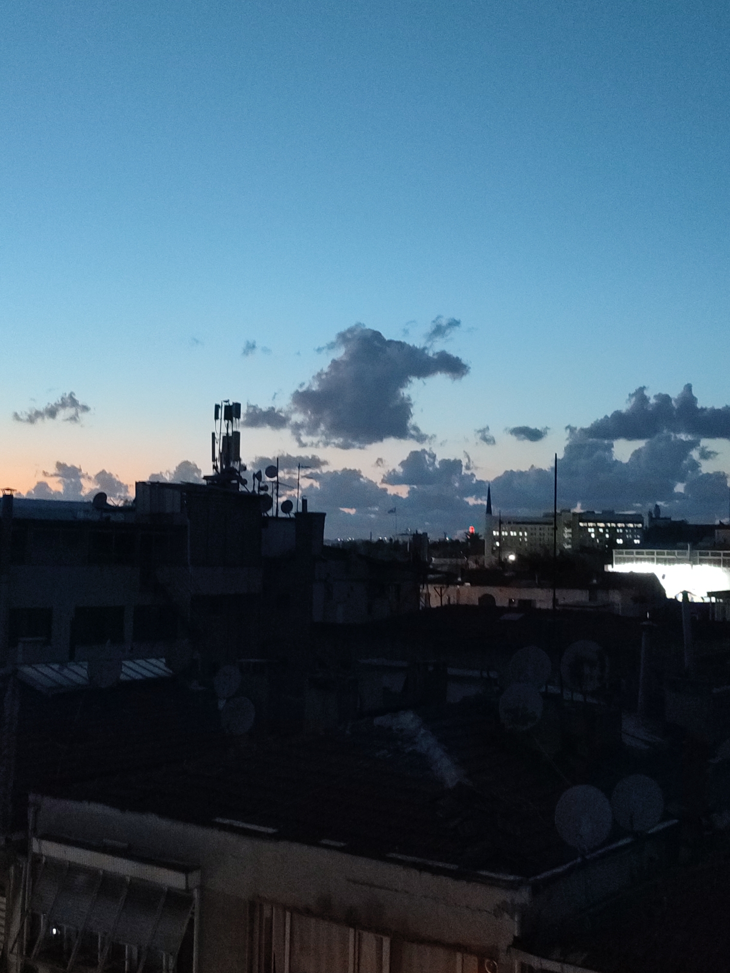 The image depicts a cityscape at dusk, likely from a rooftop in Kadıköy, Turkey. The sky is a gradient of deep blue at the top, transitioning to lighter blue and then a faint orange near the horizon, suggesting the sun has recently set or is about to rise. Scattered, dark clouds are present in the sky.
Dominating the mid-ground are the silhouetted rooftops of buildings. Several communication towers with multiple antennas are prominently visible, indicating an urban environment with a need for broadcast and mobile signals. Satellite dishes of various sizes are also scattered across the rooftops.
In the background, larger buildings are visible, some with illuminated windows, suggesting residential or commercial activity. A thin spire, possibly part of a church or a minaret, is also discernible against the sky. The foreground shows more rooftops with intricate structures and details, all rendered in dark silhouettes due to the low light conditions. The overall scene conveys a sense of urban life settling down for the night, with the remnants of daylight fading. There are no people visible in the image.