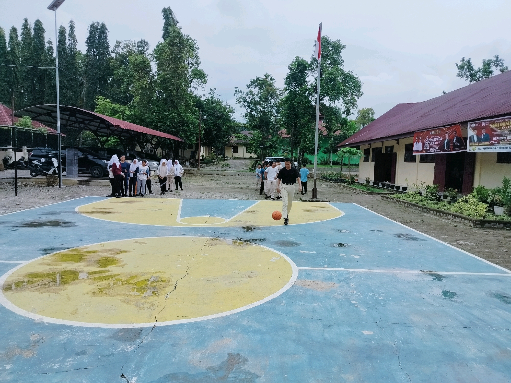 A group of young people, some wearing hijabs, stand on a gravel surface near a covered parking area with cars and a motorcycle. Adjacent to this is a basketball court, marked with blue and yellow paint and showing signs of wear and puddles from recent rain. A young man in the foreground is dribbling an orange basketball on the court. In the background, several buildings are visible, one with a red roof and banners displaying text in Indonesian. A flagpole with the Indonesian flag is also present. The environment suggests an outdoor educational or recreational facility, possibly a school campus in a moderately vegetated area under an overcast sky.