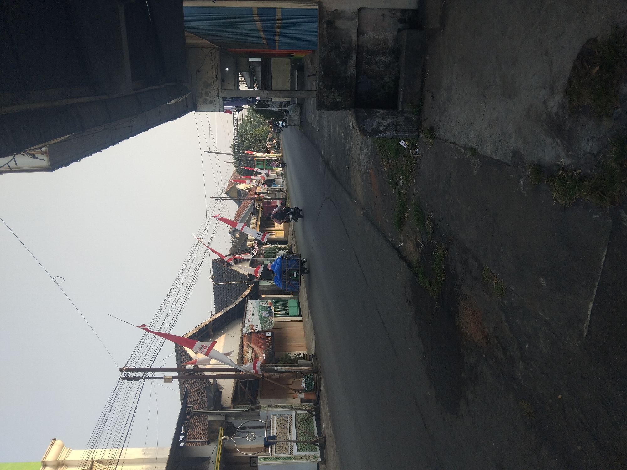 The image depicts a street scene in Tempeh, Indonesia, characterized by buildings lining both sides of a paved road under a clear sky. The architecture appears to be a mix of residential and commercial establishments, with tiled roofs and open fronts. Red and white Indonesian flags are prominently displayed along the street, suggesting a national celebration or commemoration. Several motorcycles are visible, with one carrying a passenger and another towing a cart laden with blue-covered items. A person is seen walking on the left side of the road, and another is riding a motorcycle in the distance. The road itself shows signs of wear, with some patches of discolored asphalt and scattered debris. The overall environment suggests a typical day in a bustling Indonesian town, with daily activities and local decorations present.