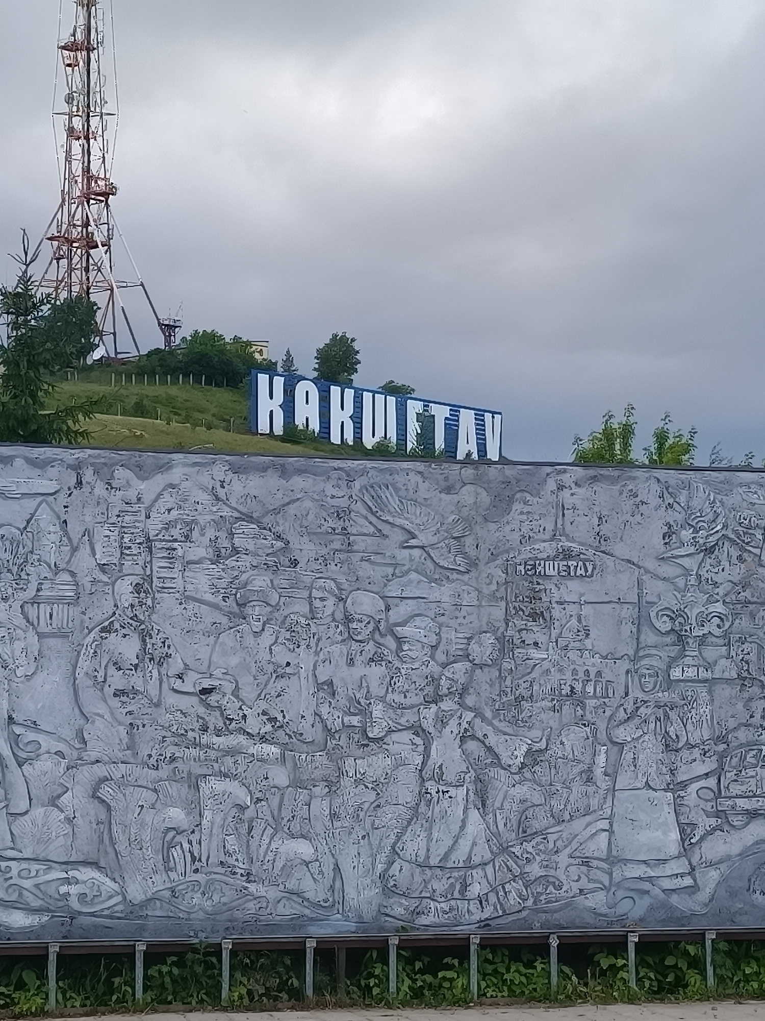 The image shows a monument with a relief depicting historical scenes in Kokshetau, Kazakhstan. Above the monument, on a hillside, are large white letters spelling out "KOKSHETAU". A tall, red and white communication tower rises in the background. The sky is overcast. Below the monument is a small strip of greenery.