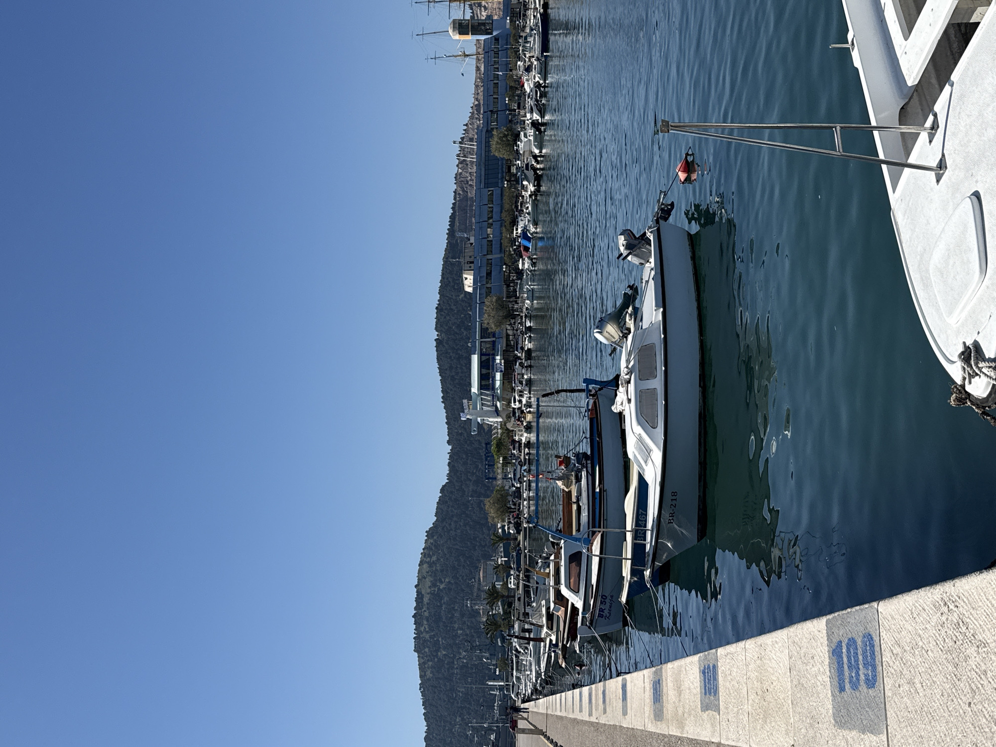 The image captures a marina in Bar, Montenegro, lined with various boats docked along a concrete pier. Clear blue sky dominates the background. Boats of varying sizes and types are moored; one visible boat has the registration number "BR-218". The pier is marked with blue-painted numbers, including "109". A tree-covered mountain range is visible in the distance behind the marina. The water is calm, reflecting the surrounding environment.