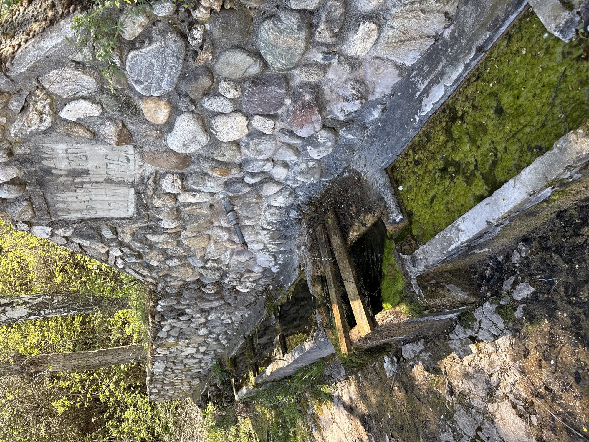 The image depicts a stone structure likely related to a water system, possibly a series of connected reservoirs or channels. The structure is constructed from irregularly shaped stones and concrete. Some sections of the channels are filled with green algae-covered water. Wooden beams are present, possibly for support or division. A stone plaque with indistinct markings is integrated into the stone wall. Vegetation is visible at the periphery of the structure. The location is Boboshevo, Bulgaria.