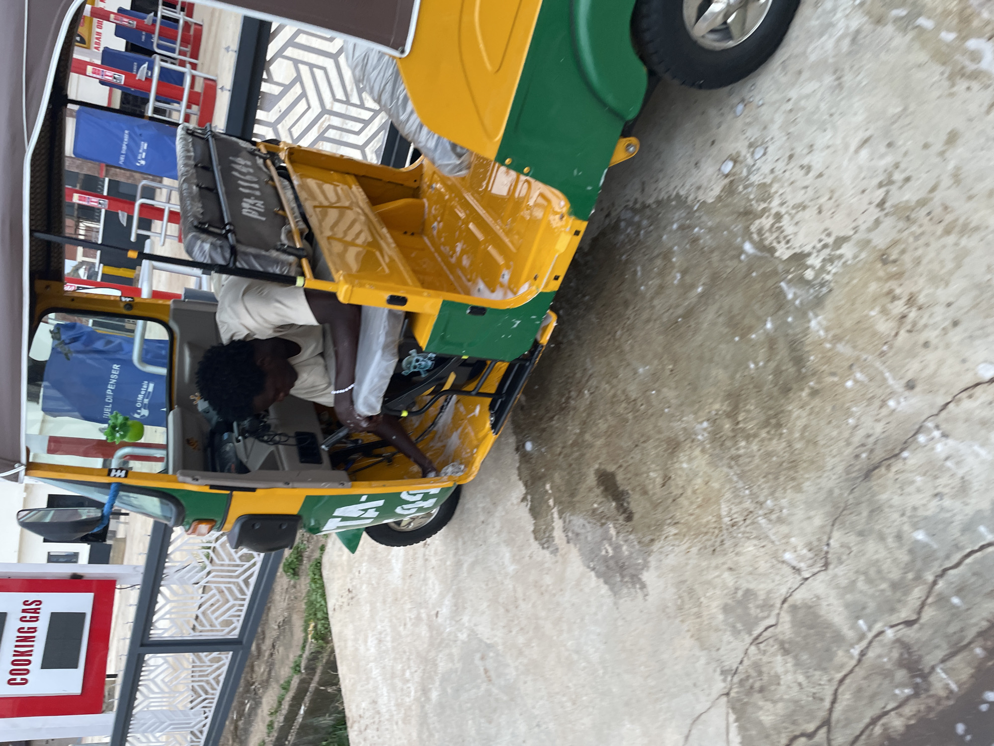 In Barakin Lerawi, Nigeria, a young man, appearing to be in his late teens or early twenties, is inside the driver's seat of a yellow and green auto-rickshaw. He is wearing a light-colored t-shirt and has a necklace or bracelet on his left wrist. He is bent over and appears to be cleaning or attending to something within the vehicle's interior, possibly the floor area. The auto-rickshaw has a covered roof and a passenger seating area behind the driver. In the background, there are fuel dispensers, suggesting the scene is at a gas station. The ground is paved with concrete and shows signs of wetness, with some soapy suds near the auto-rickshaw, indicating it may be undergoing a wash. A white railing with an abstract pattern is visible to the left of the auto-rickshaw. A red sign with white lettering reading "COOKING GAS" is also visible on the left side of the image.