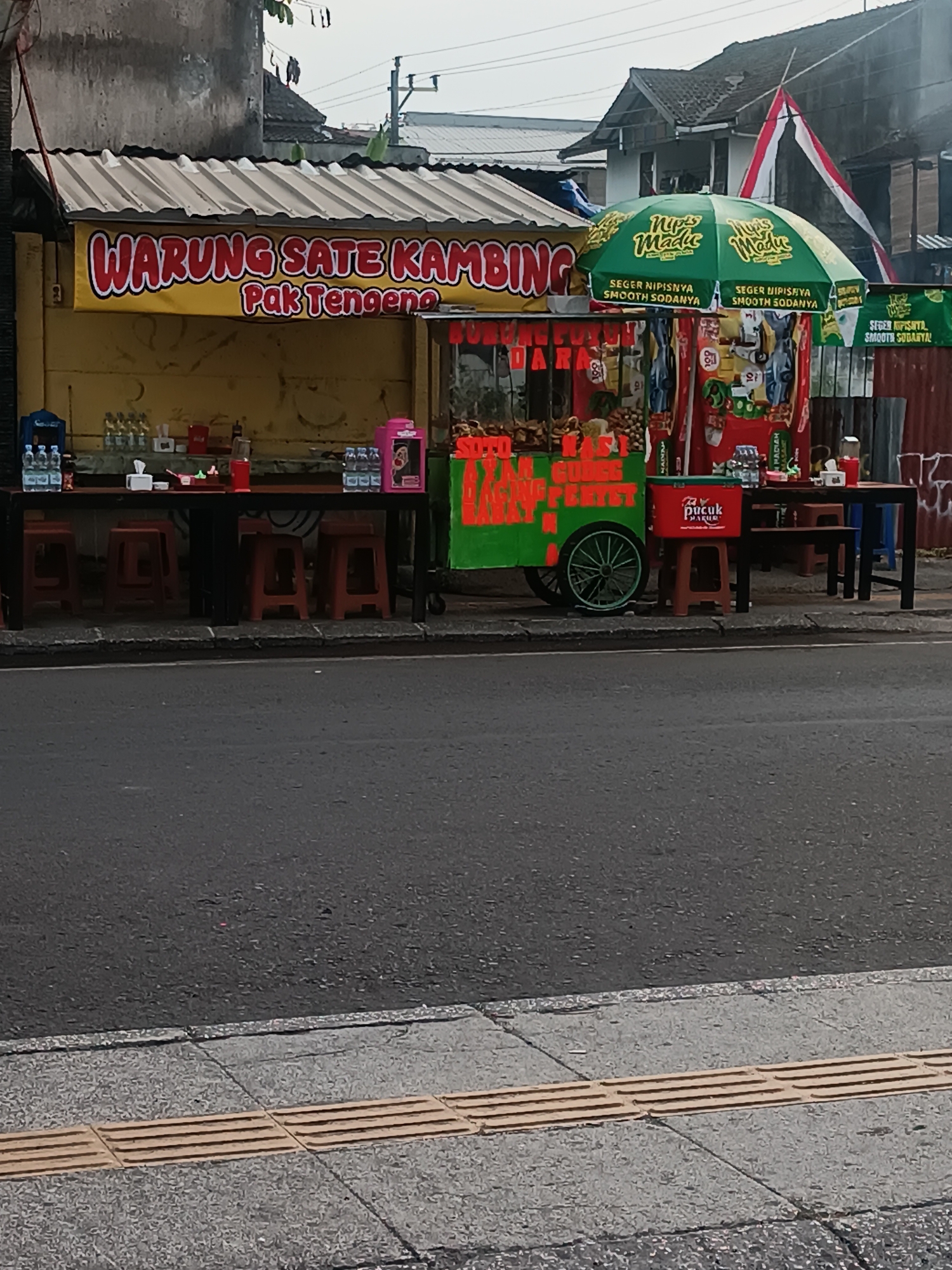 The image captures a street-side food stall in Yogyakarta, Indonesia, likely in a moderately urban setting. A prominent yellow awning displays the text "WARUNG SATE KAMBING PAK TENGENG" in red and white lettering, indicating it is a goat satay restaurant. In front of the stall, a long dark table is set up with red plastic stools, suggesting seating for customers. Various items, including bottled drinks and tissue boxes, are arranged on the table.
To the right of the main stall, there is a smaller cart, possibly a mobile beverage or snack vendor, adorned with colorful graphics and featuring a green umbrella with the logo "Nups Madu". This umbrella also displays the text "SEGER NIPISNYA SMOOTH SODANYA", advertising a fresh and fizzy soda. Behind the stalls, residential buildings with tiled roofs are visible, and a red and white Indonesian flag is draped from one of them. The street in the foreground is paved with asphalt, and a textured sidewalk with raised tactile paving is present on the right side of the image, typically found in pedestrian areas. The overall scene depicts a typical day at a local eatery in Yogyakarta.