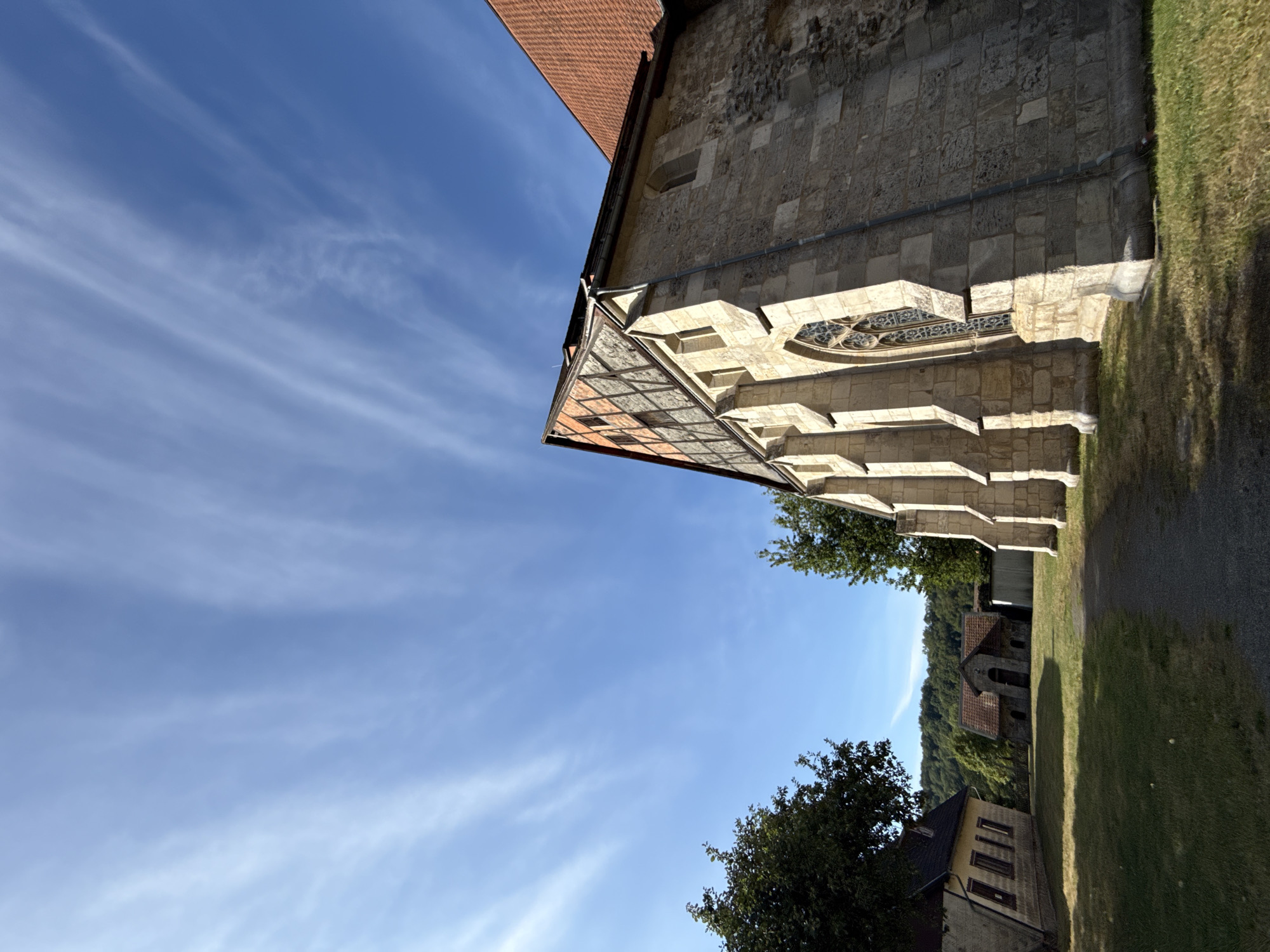 The image depicts an exterior view of the historic St. Nicholas' Church in Walkenried, Germany. The church, constructed of stone with a red-tiled roof, features arched windows and buttresses along its side. Adjacent to the church are other stone buildings with tiled roofs, resembling historical outbuildings or residences. The scene is set on a grassy area with a gravel path leading towards the church. Lush green trees are present on the left and right sides of the frame, and a wooded hillside forms the background. The sky is a clear blue with thin, wispy white clouds, indicating a daytime setting under fair weather conditions. The perspective is low-angle, emphasizing the scale of the church. No people are visible in the image.