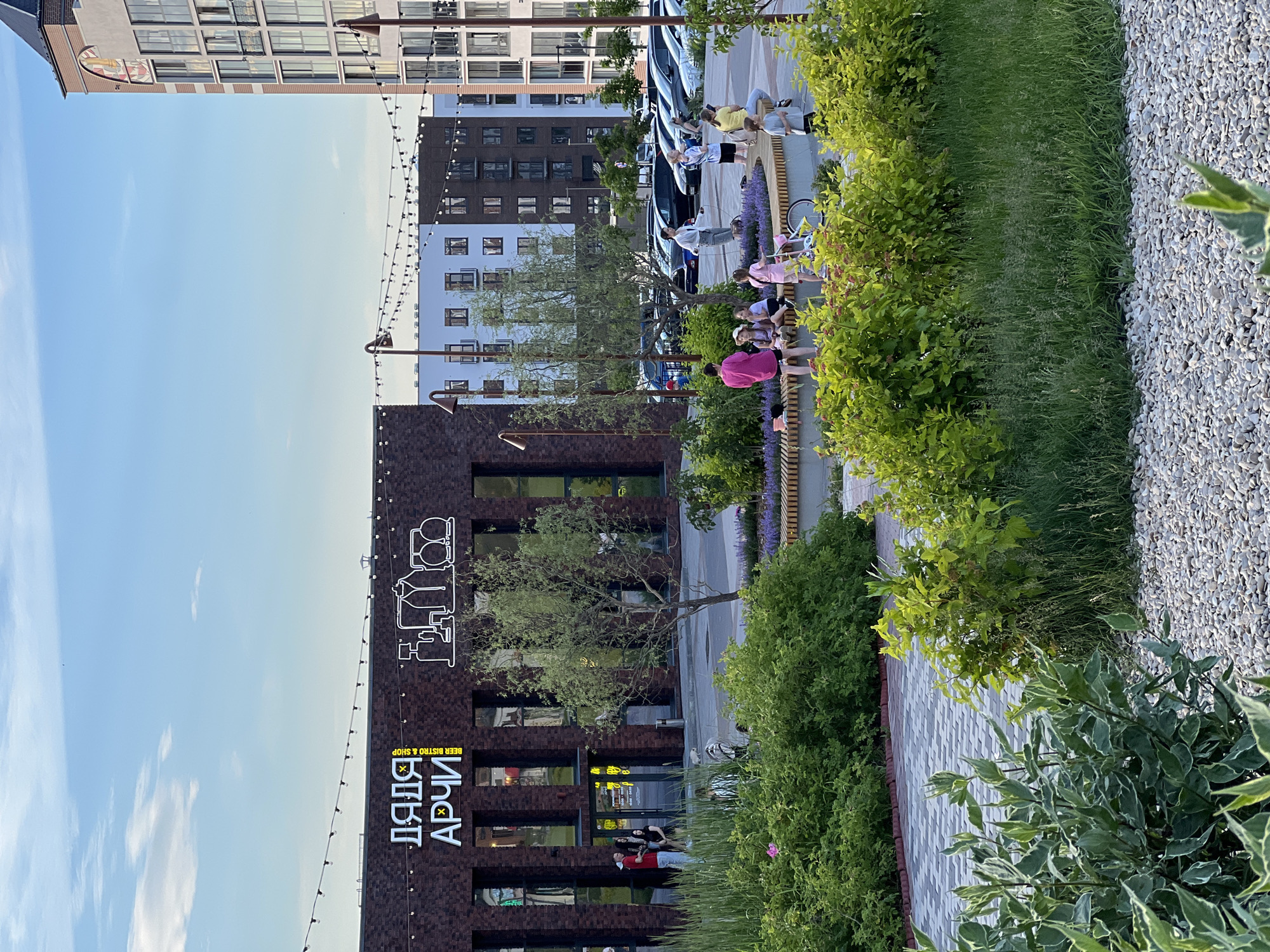 The image depicts an outdoor scene with a building and surrounding landscaping. A brick building with large windows is visible, featuring a prominent neon sign with Cyrillic text that reads "ДЯДЯ АРЧИ" and "BEER BISTRO & SHOP." In front of the building, several people are gathered on a bench and standing. The individuals appear to be casually interacting, with some sitting and others standing. The setting includes lush green bushes and trees, a paved walkway with a patterned design, and a gravel area. String lights are strung across the scene, suggesting it might be an evening or a festive atmosphere. The sky is a clear blue with some wispy clouds, indicating fair weather and likely daytime. The overall impression is a modern urban space with a focus on leisure and community gathering. The presence of the beer bistro sign and people relaxing suggests a relaxed and social environment. The architecture of the building, with its contemporary design and large windows, points to a modern urban development.
