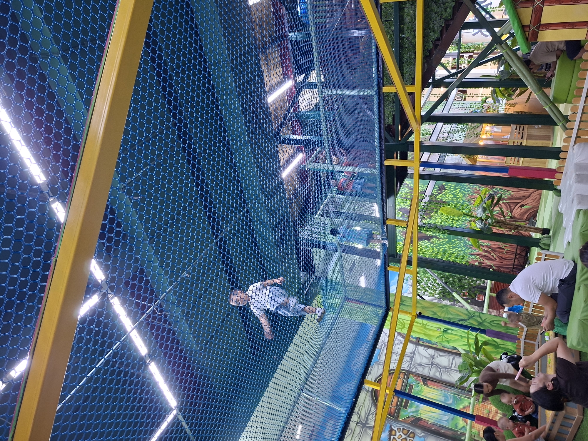 The image depicts an indoor children's play area, likely a soft play center or trampoline park, with a jungle-themed decor. A young boy in a white t-shirt and shorts is captured mid-air, jumping on a trampoline surface enclosed by blue netting. The structure features yellow metal beams and supports, with further levels visible in the background. In the lower part of the image, several adults and children are gathered, with some seated on green cushions. The walls are decorated with large, colorful murals of trees and foliage, creating an immersive environment. The lighting appears to be artificial, with bright fluorescent tubes visible on the ceiling. While the exact location within Bishkek City, Kyrgyzstan, is not specified, the scene suggests a family-friendly entertainment venue.