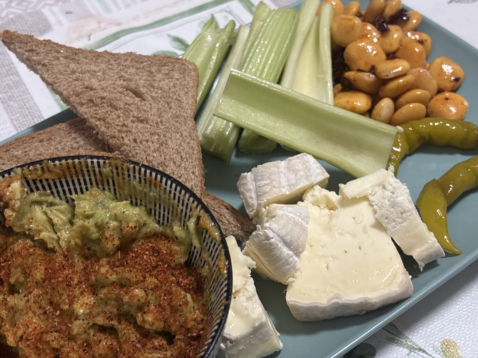 A close-up, high-angle shot shows a light teal plate filled with a variety of food items, placed on a table covered with a white tablecloth featuring a subtle green floral pattern. In the foreground to the left, a striped ceramic bowl contains a chunky, light green dip, possibly avocado, generously sprinkled with a reddish spice mix. Next to the bowl, several triangular slices of whole wheat toast are arranged.
The plate itself holds several elements: a cluster of golden-brown beans, possibly butter beans or lima beans, coated in a glistening sauce with visible dark pieces, likely dried fruit or herbs. To the right of the beans are two bright green pickled peppers. Several stalks of crisp, pale green celery are neatly arranged on the plate. In the lower right section of the plate, several wedges of white cheese, some with rinds, are visible. The overall presentation suggests a casual appetizer or snack spread. The image does not contain any visual cues to identify a specific location in Barcelona, Spain.