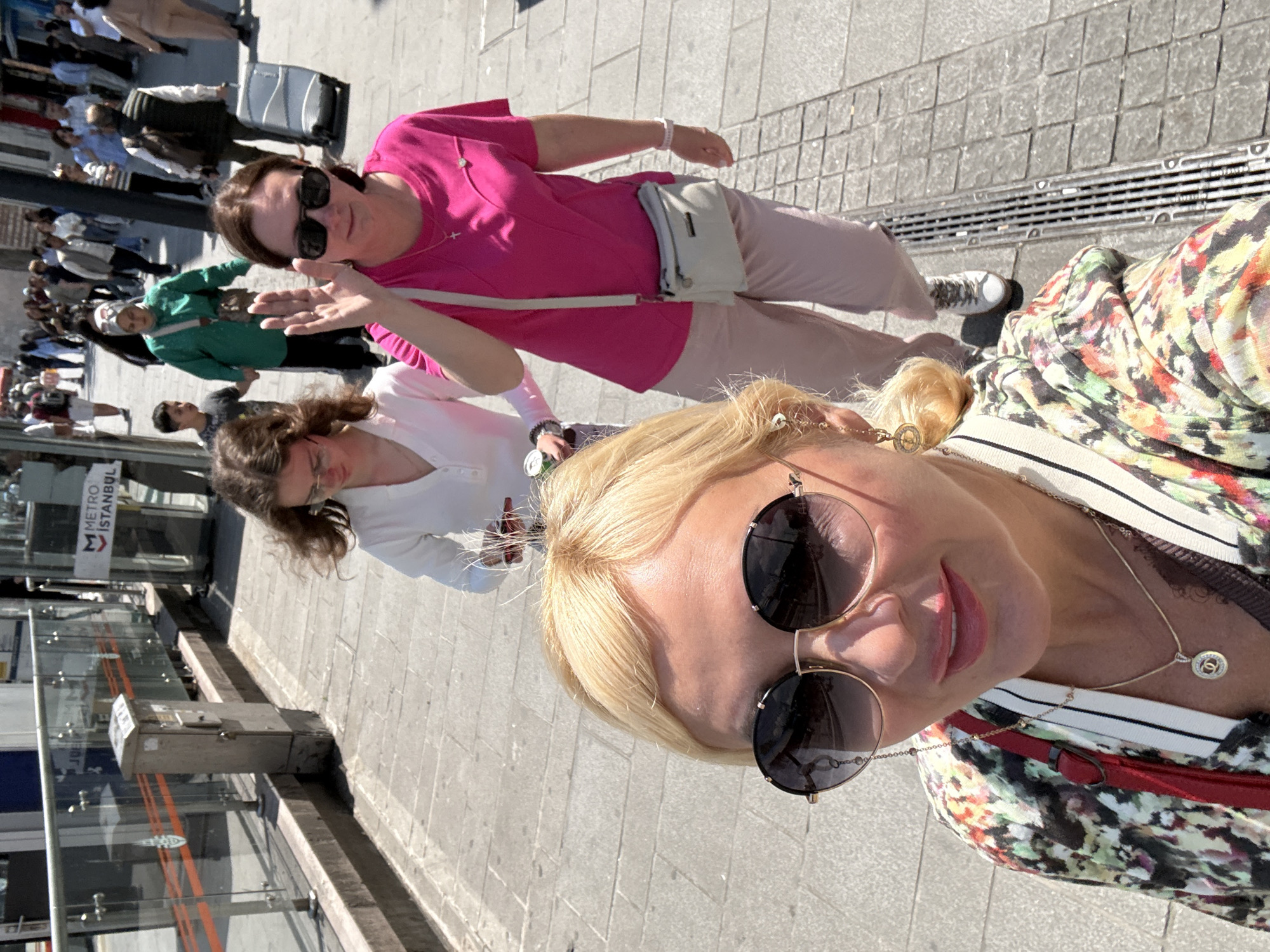 The image shows four women standing outside a Metro Istanbul station. The foreground shows a blonde woman wearing sunglasses and a colorful shirt, suggesting a selfie. Behind her are two other women; one wearing a pink shirt is waving while the other is wearing a white shirt. In the background, a crowd of people is visible, along with the entrance to the Metro Istanbul station. The setting appears to be a sunny day on a city street.