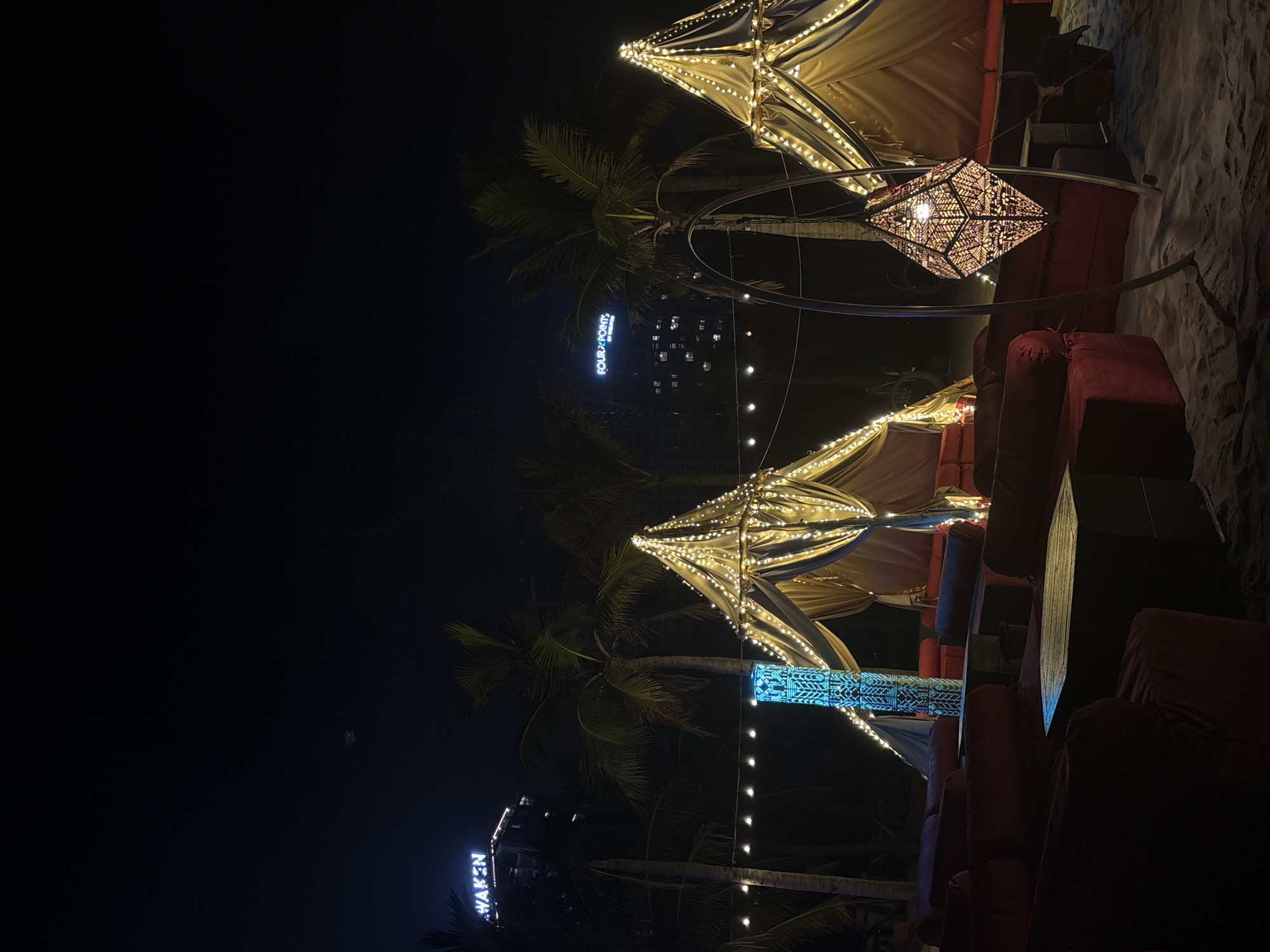 The image shows an outdoor nighttime scene, possibly a beachside lounge or bar in Đà Nẵng, Vietnam. There are cushioned seating areas, illuminated by string lights and decorative lamps. The overall ambiance appears upscale and relaxed. Palm trees are visible in the background, along with a building with illuminated signs "WAKEN" and "FOUR POINTS BY SHERATON."