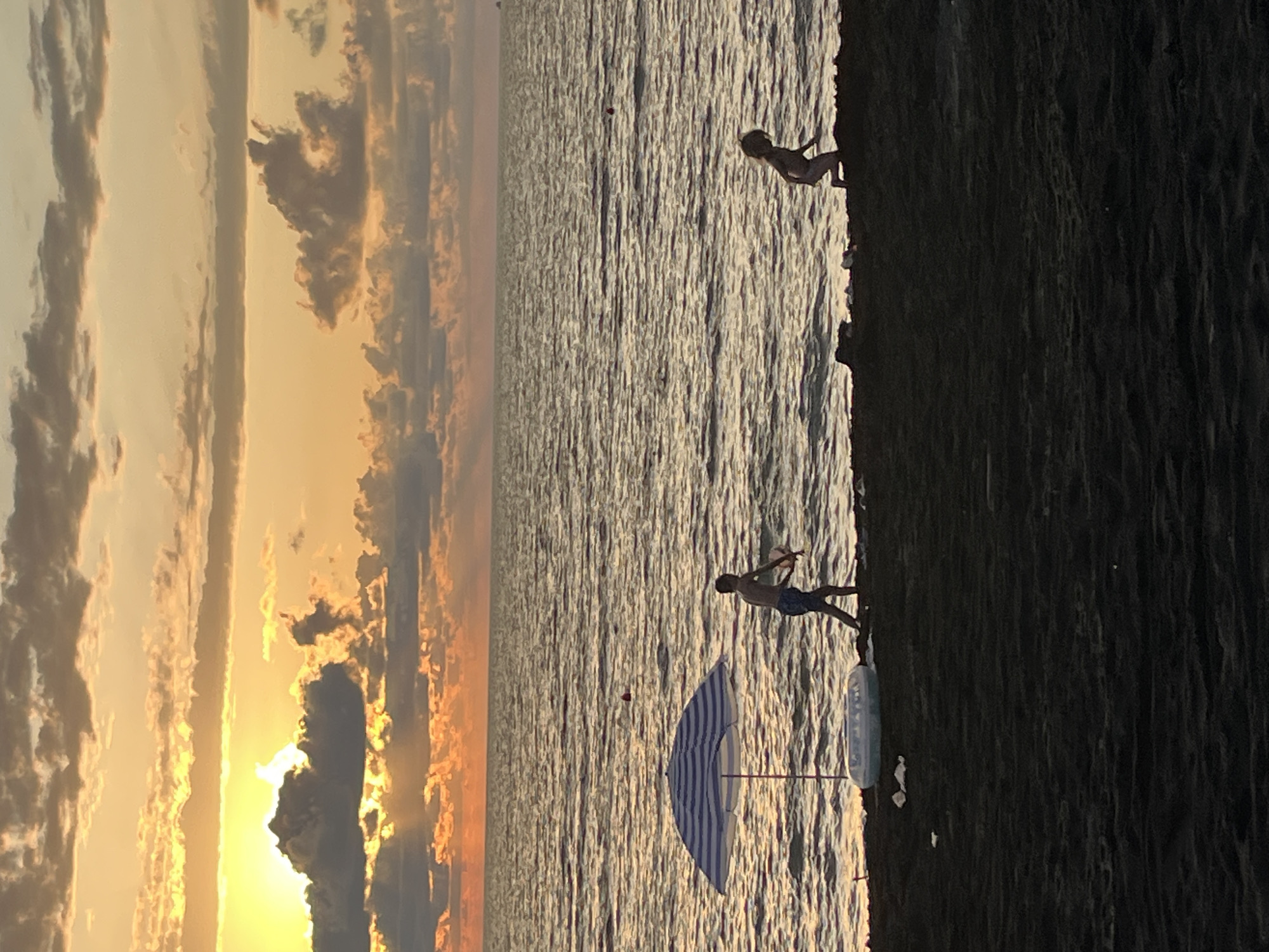 This image depicts a sunset scene at the beach in Ureki, Georgia. The sky is filled with dramatic clouds illuminated by the setting sun, casting a warm, orange glow. The sea is calm with gentle waves. In the foreground, a dark sand beach is visible.
Two children are present on the beach. A boy, silhouetted against the water, is standing and appears to be playing with a toy. To his right, a smaller child, also silhouetted, is in a kneeling or crouched position. A blue and white striped beach umbrella is set up on the left side of the image, next to an inflatable pool or raft. The overall atmosphere is serene, capturing a moment of relaxation and play by the sea at dusk.