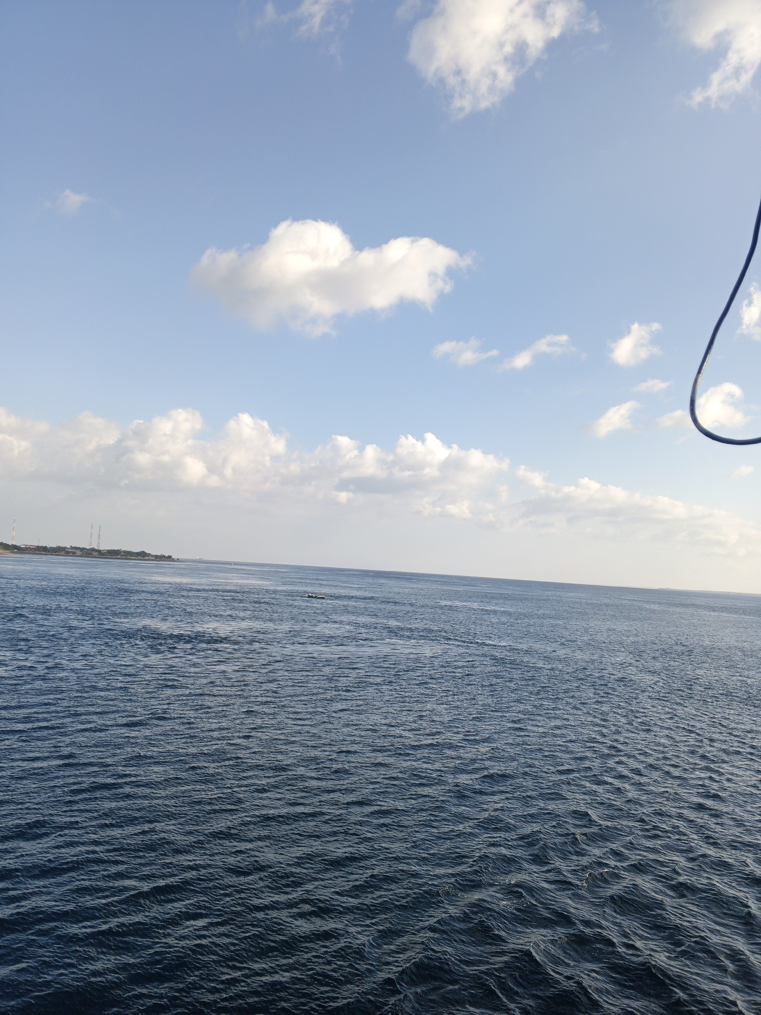 The image shows a wide expanse of deep blue water with gentle ripples under a bright blue sky dotted with fluffy white clouds. In the distance, a thin strip of land is visible, lined with structures including what appear to be radio towers. The coastline suggests a developed area, possibly a port or industrial zone. A small, dark boat with two occupants is visible on the water to the right of the center. A black, curved cable hangs from the upper right corner of the frame, indicating the viewpoint is likely from a boat or elevated structure. The overall impression is a serene maritime scene, likely captured on a clear day in the vicinity of Melaya, Indonesia, given the coastal landscape and tropical cloud formations.