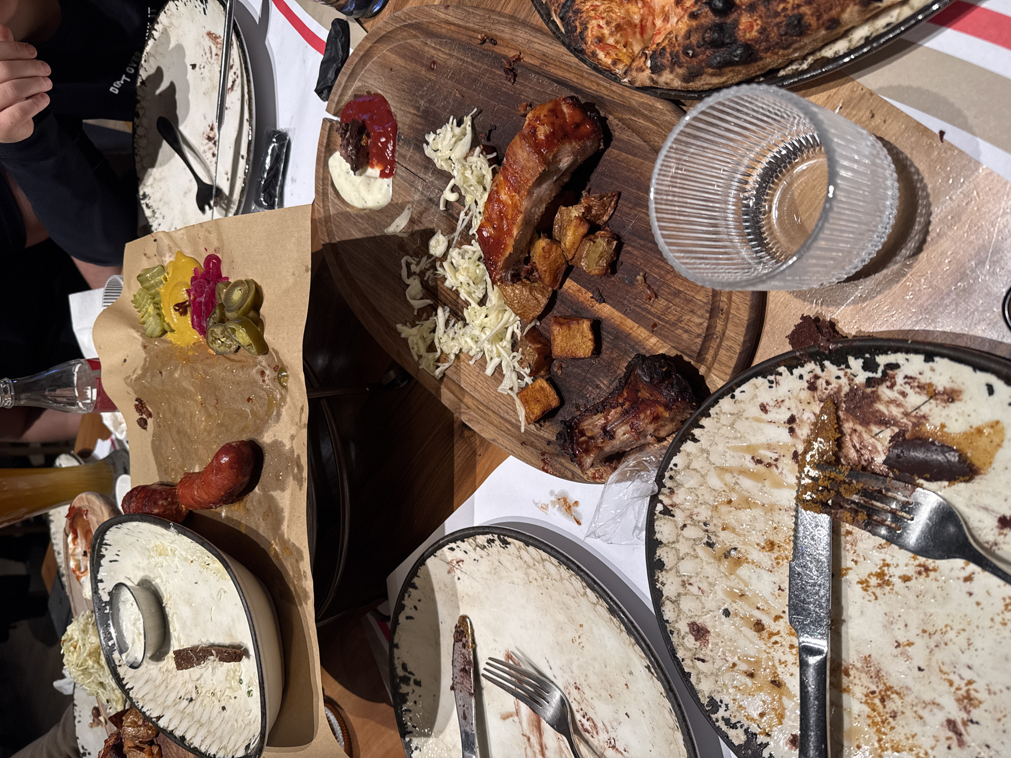 The image depicts a table in what appears to be a restaurant in Yerevan, Armenia, covered with remnants of a meal. Several partially-eaten plates are scattered across the surface, containing leftover food and used cutlery. On a wooden board, a piece of rib is visible alongside roasted potatoes and coleslaw. Next to the board, an upside-down empty glass sits. The table also holds a paper wrapping with pickles, and sliced sausage. In the background, parts of a pizza are visible. A person's arm is seen on the left, suggesting a casual, social dining atmosphere. The scene suggests a meal is completed.