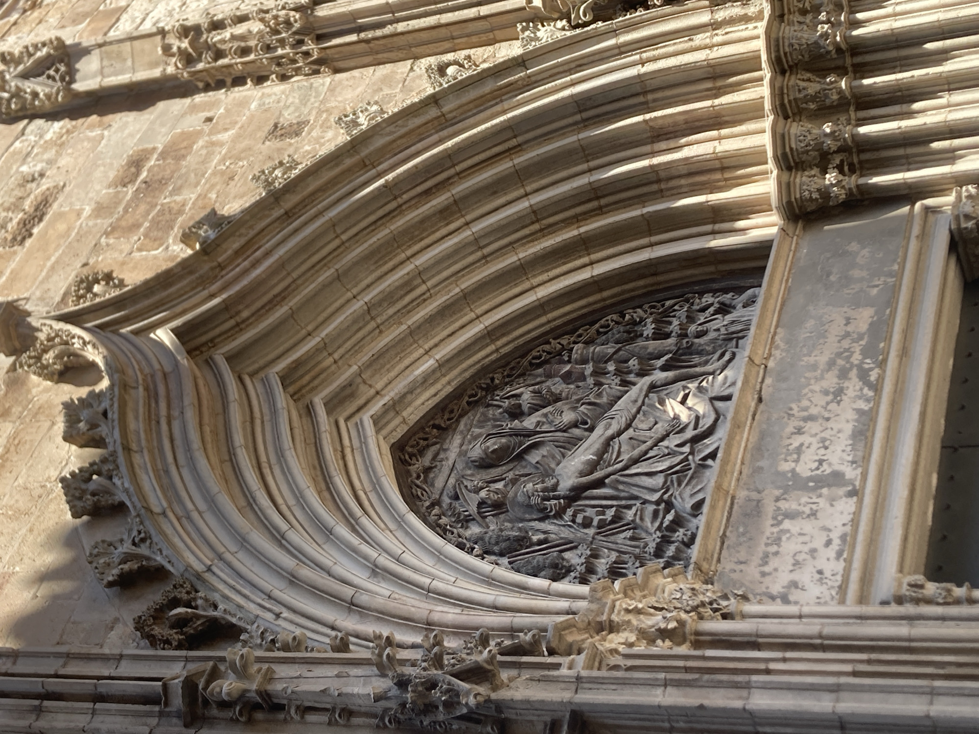 This is a close-up, eye-level, exterior shot of a Gothic cathedral entrance in Barcelona, Spain. The focus is on a detailed stone relief carving above an arched doorway. The relief depicts a scene from Christian scripture, likely the Pietà, with a central figure of Mary holding the body of Jesus. Several other figures are present, suggesting a larger narrative within the relief. The stone archway above the relief is elaborately carved with multiple ribbed layers, characteristic of Gothic architecture. The surrounding stonework of the cathedral facade is weathered and shows the texture of aged stone blocks. Sunlight casts strong shadows, highlighting the three-dimensional details of the carvings. The image captures the intricate artistry and monumental scale of religious architecture.