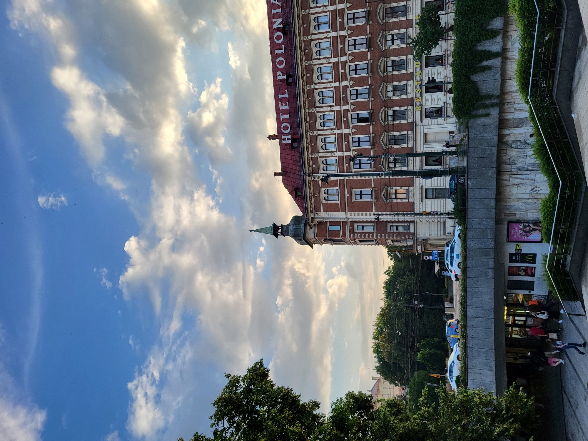 This is a vertical, low-angle, outdoor photograph taken from a street level perspective looking up towards a building. The building is made of red brick and has many windows. The words "HOTEL POLONIA" are visible in large red letters across the top of the building. The sky is blue with white, fluffy clouds. There are trees in the foreground and middle ground. The scene appears to be a city street. There are a few people visible, walking along the sidewalk in front of the building. There is also a white car parked on the street. The lighting suggests it is daytime, possibly late afternoon or early evening.