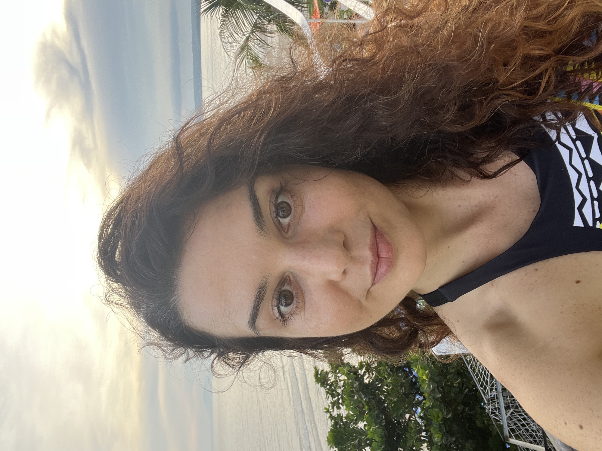 A woman with long, wavy brown hair is taking a selfie outdoors. She is wearing a black swimsuit with a colorful, geometric pattern. Her head is turned slightly to her left, and she is smiling. In the background, there is a body of water, possibly the ocean, with gentle waves. Palm trees and lush green foliage are visible on the left side of the image, suggesting a tropical or subtropical location. The sky is overcast with diffused sunlight, creating a soft, bright atmosphere. The setting appears to be Mũi Né, Vietnam, a coastal city known for its beaches and resorts.