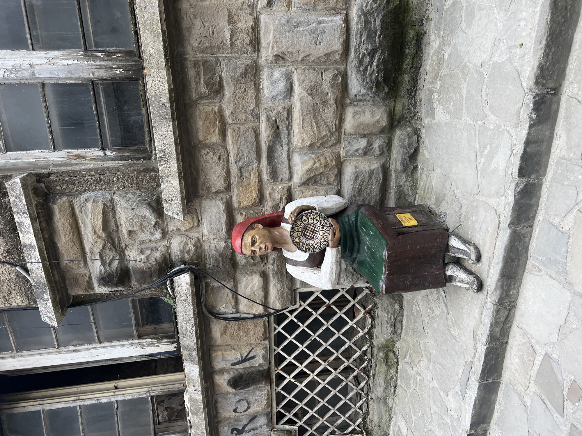 This image, set in Veliko Tarnovo, Bulgaria, captures a rustic, colorful statue of a woman positioned against a historical stone building. The statue, depicting a woman in traditional attire, wears a red headscarf, a white blouse or apron, a green skirt, and grey shoes. She holds a decorative circular plate with a prominent black and yellow/gold mandala-like pattern, as if presenting it.
The statue stands on a brown, box-like base. To the left, an old, multi-pane window with peeling white paint on its wooden frames is visible, its glass appearing dark and reflective. Below the statue, a white, diamond-patterned metal grille covers an opening in the stone wall. Black utility cables run horizontally across the textured stone, passing near the statue. The building's walls are constructed from irregular, rough-hewn stone blocks in shades of light grey and tan. The ground or adjacent pathway features lighter, irregularly shaped flagstones. Faint graffiti, including the letters "RK," can be seen on the stone wall below the window. A small, yellow label with illegible text is attached to the statue's brown base. The scene appears to be under natural daylight, possibly overcast, contributing to diffused lighting and soft shadows.