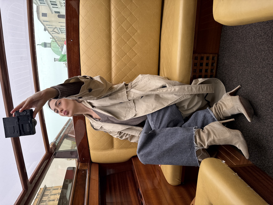 A young adult female is depicted taking a selfie inside a water taxi in Venice, Italy. She is seated on a golden-yellow, diamond-quilted upholstered bench, dressed in a beige trench coat, a brown V-neck sweater, dark blue wide-leg jeans with frayed hems, and light beige suede high-heeled ankle boots. Her right arm is extended, holding a black digital camera with a flip-out screen pointed towards herself, while her lips are puckered in a selfie pose. The boat's interior features polished wooden trim and a dark grey carpeted floor. Through the boat's windows, Venetian architecture is visible under an overcast sky, including a prominent green dome, consistent with the dome of San Geremia, and a partially visible Italian flag.