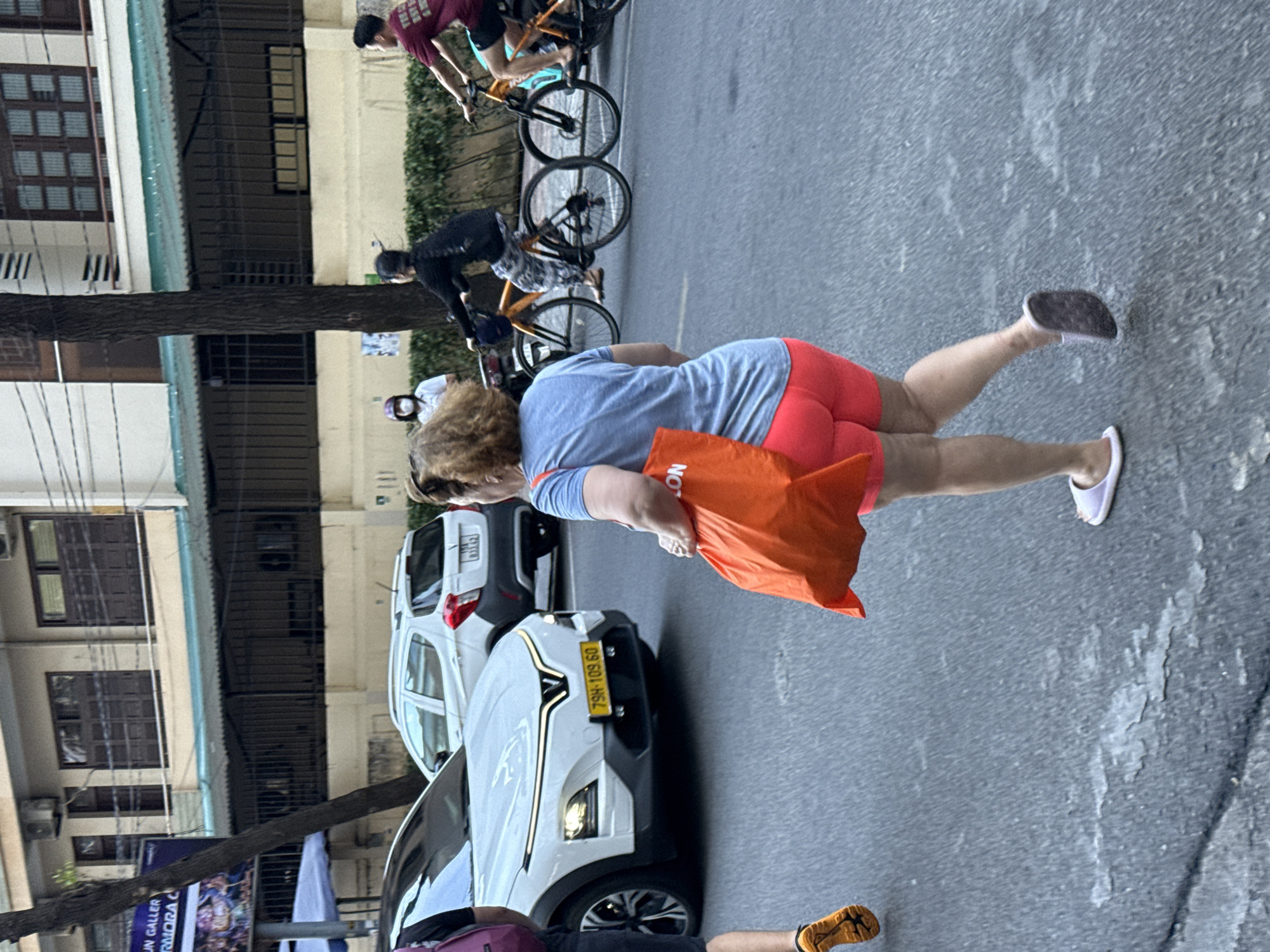 The image depicts a street scene in Nha Trang Ward, Vietnam, with several people and vehicles present. In the foreground, a woman with curly blonde hair is walking, holding an orange bag. She is wearing a blue t-shirt and red shorts. Her right leg is extended as if she is in mid-stride, and she is wearing white slip-on shoes. Her left leg is bent, and she is wearing a dark shoe.
Behind her, a white car is parked, with its front end facing the camera. A yellow license plate is visible on the car, reading "79H-103.60". Another white vehicle is partially visible behind the first car.
In the background, several people on bicycles are visible. One person is wearing a maroon shirt and another is in dark clothing. The bicycles have multiple gears and are positioned on the street. Buildings line the street, with windows and air conditioning units visible on the facade of one building. A tree trunk is prominent in the middle ground, casting a shadow. A poster with some text is visible on a fence in the background.
The scene appears to be taking place during the daytime, likely in an urban or suburban area. The lighting suggests it might be overcast or late afternoon, given the shadows. The overall impression is a busy street with everyday activity.