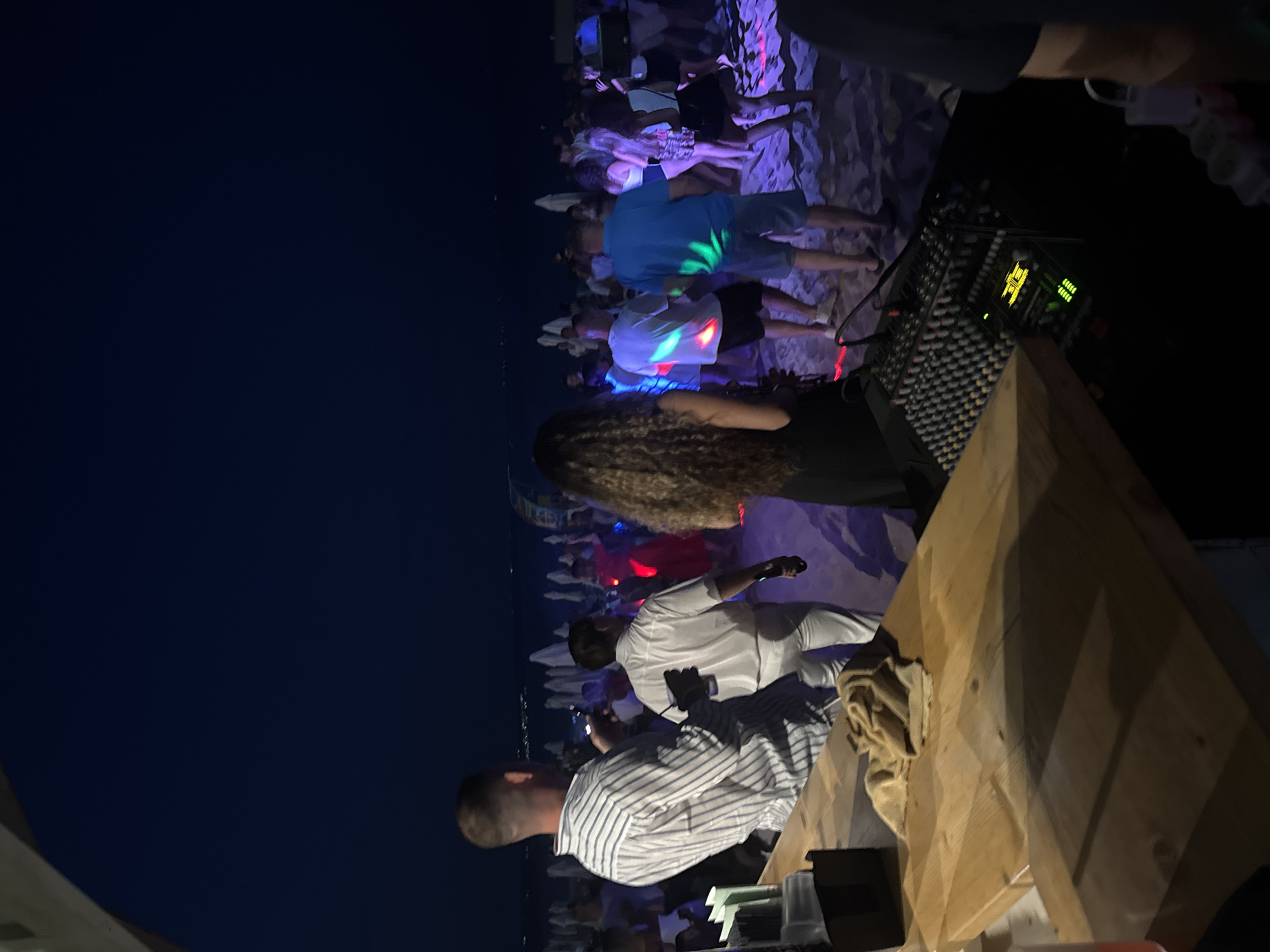 An evening outdoor party is underway on a sandy beach in Varna, Bulgaria. In the foreground, a wooden bar counter is visible, with a sound mixer placed on it, its lights indicating activity. Behind the bar, a man in a striped shirt is observing the scene, while another man in white attire walks away from the camera. Further back, a crowd of people are dancing on the beach, illuminated by colorful lights from above and from the ground. Beach umbrellas are set up in the background, suggesting a resort or organized event. The dark sky overhead and the distant lights on the horizon indicate nighttime in the city.