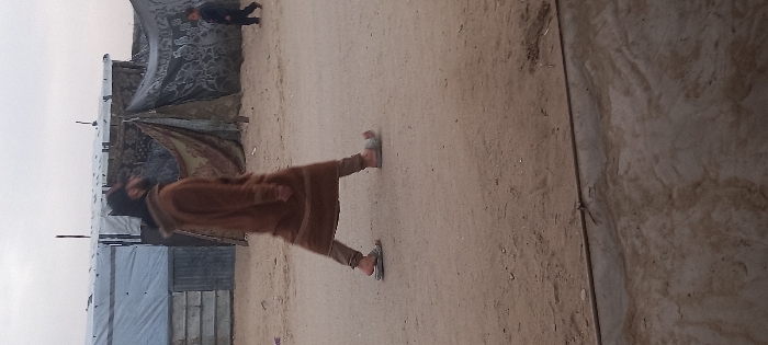 An outdoor scene in Khan Yunis, Palestinian Territory, depicts a dry, sandy ground under an overcast sky. In the mid-ground, a bearded adult male walks from right to left, wearing a brown hooded garment, light-colored trousers, and grey open-toed sandals. His head is turned partially to the left. In the background, further to the right, a small child dressed in dark clothing also walks from right to left. Behind the individuals, a series of makeshift structures are visible. To the left, a structure with light-colored walls and a metallic or tarp roof stands next to another tent-like dwelling made of patterned fabric. Adjacent to this, a darker tent-like structure is also visible. The ground in the foreground is unpaved, with a distinct horizontal line marking a change in surface texture at the very bottom of the frame.