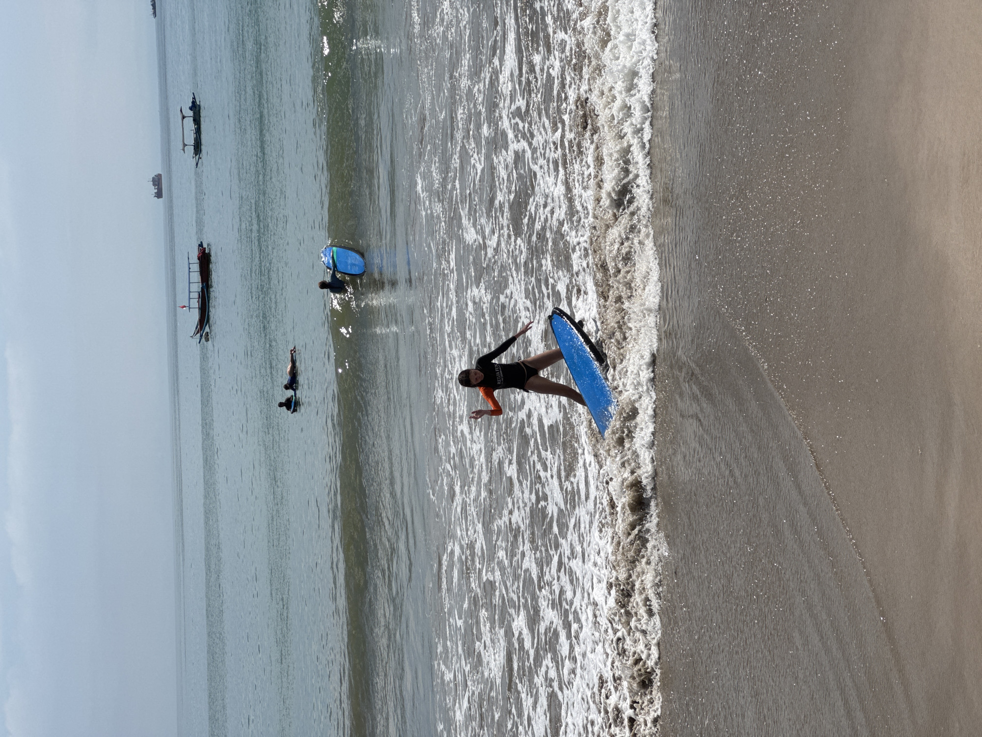 The image depicts a beach scene in Kuta Selatan, Indonesia. In the foreground, a person in a black wetsuit with orange sleeves stands in the shallow surf, holding a blue surfboard. The person faces towards the ocean.
Further out, two other individuals are visible in the water with surfboards, one blue. Several small fishing boats dot the horizon. The sky is overcast, creating a muted light across the water and sand. The beach itself is sandy, and the waves are small.