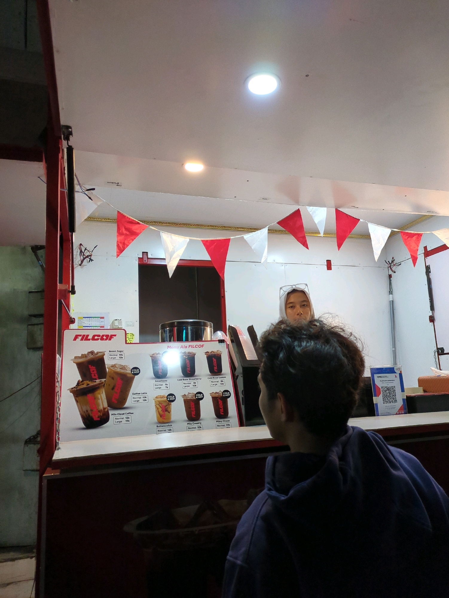The image shows an outdoor food stall in Gumpang, Indonesia. The stall has a red and white triangular banner strung across the top of the counter. There is a menu board on the left displaying various drinks with prices, and to the right is a woman wearing glasses and a headscarf, presumably the vendor, standing behind the counter. In the foreground, a person wearing a purple hoodie is facing away from the camera, looking at the menu board. The stall appears to be open for business at night, as indicated by the artificial lighting.