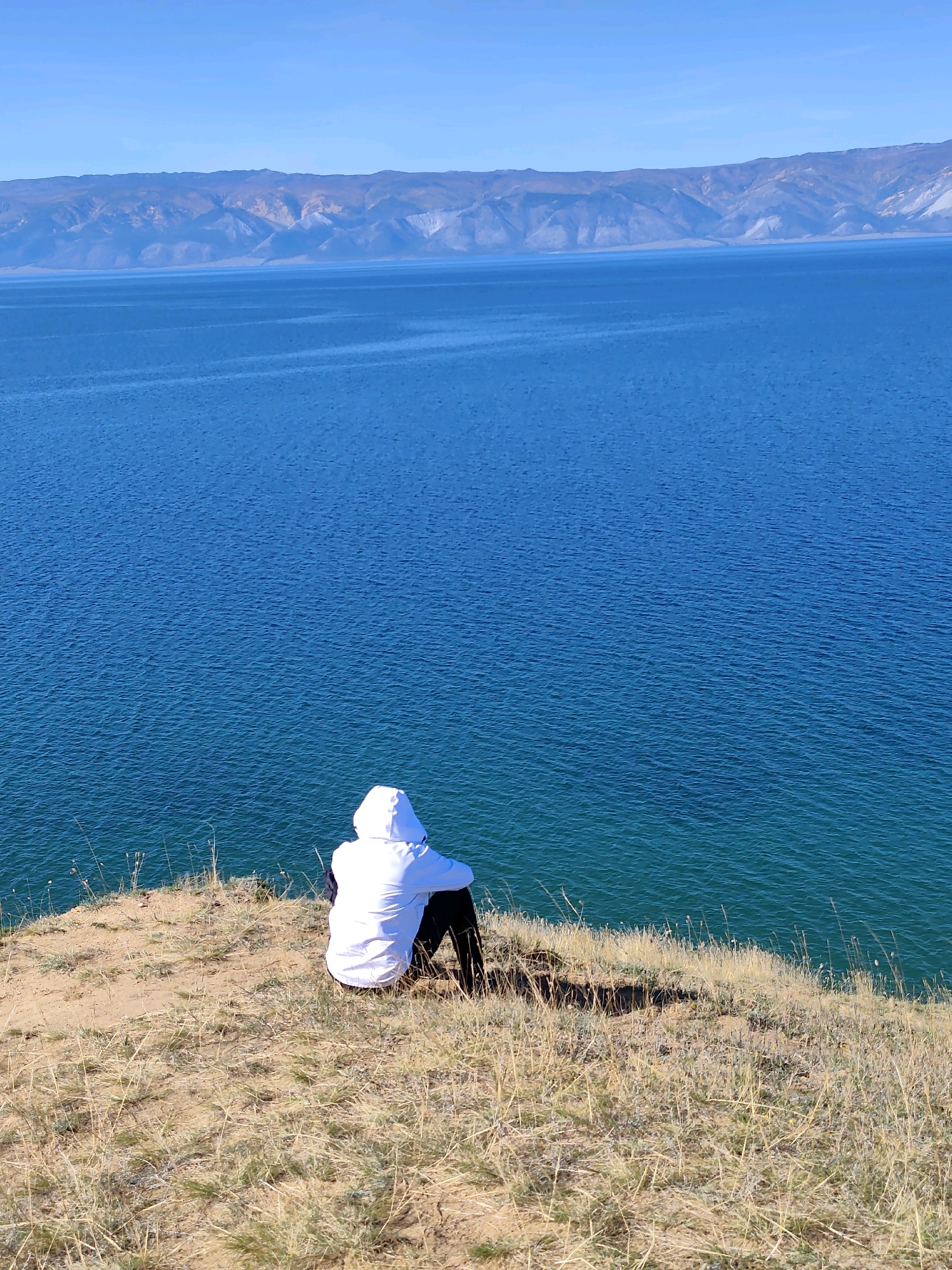 A person wearing a white hooded jacket and dark pants sits on a grassy cliff overlooking a large body of deep blue water. The water stretches to the horizon, where distant, hazy mountains can be seen under a clear, pale blue sky. The grassy area in the foreground is dry and sparse, with patches of exposed earth. The scene appears to be in a natural, open environment, likely on the shore of Lake Baikal near the village of Kharantsy, Russia, given the distinctive landscape. The person is facing away from the camera, looking out at the water.