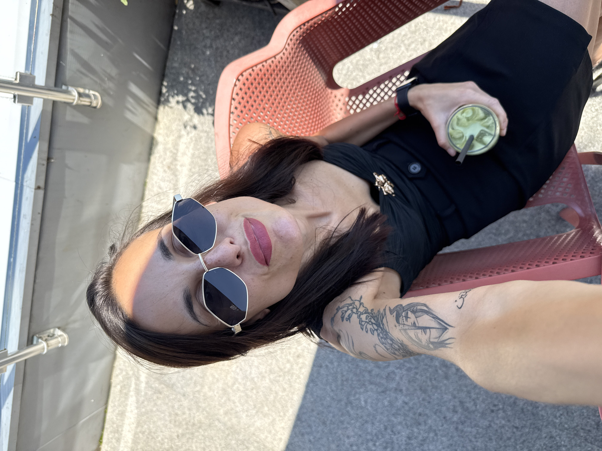 A woman is lying on a red plastic chair outdoors. She is wearing sunglasses and a black dress. Her left arm has a tattoo and she is holding a green beverage with a straw. The background suggests an urban environment, possibly in Pleven, Bulgaria, given the prompt's information. The scene appears to be a casual, relaxed setting.