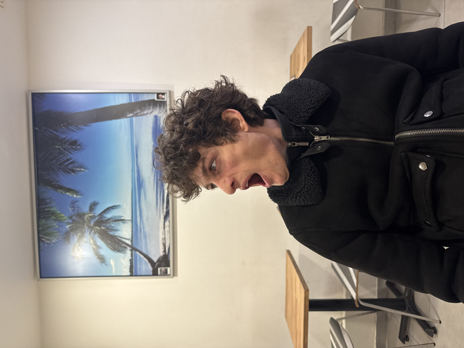 The image depicts a young adult male seated indoors, identified as being within the Frigo 27 burger restaurant located at 27 Rue Sainte-Croix de la Bretonnerie in the Marais district of Paris, France. The male subject, possessing curly dark brown hair and light-colored eyes, is shown from the chest up, oriented with his body slightly angled and his head sharply turned to his right (viewer's left). He wears a black jacket with a faux shearling collar, a prominent silver zipper closure, and two flap pockets secured by silver snap buttons on the lower front. His mouth is wide open, revealing his teeth and tongue, as if caught in a moment of vocalization, surprise, or a wide yawn, with his gaze directed off-camera to his right.
The background features a plain white wall. Centered behind the subject is a large, rectangular framed picture displaying a vibrant tropical beach scene with a bright blue sky, a turquoise ocean, and two prominent palm trees. The bottom left corner of this frame includes a small white square with text, clearly showing "FRIGO 27" and "BOITE," which serves as the identifying detail for the location. Partially visible to the left and right of the subject are small, square wooden tables with dark metal pedestal bases. A chair with a distinctive back featuring horizontal white and dark grey stripes is partially visible to the left, positioned under one of the tables.