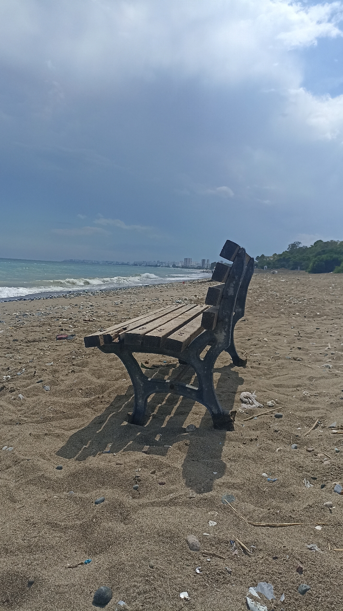 An empty, weathered wooden bench with dark metal legs sits on a sandy beach in Mezitli, Turkey. The bench is angled to the right, facing the sea. The sea has moderate waves and a coastline with buildings and trees in the distance. The sky is partly cloudy. The sand is littered with small stones, shells, and debris. The shadow of the bench is cast on the sand.