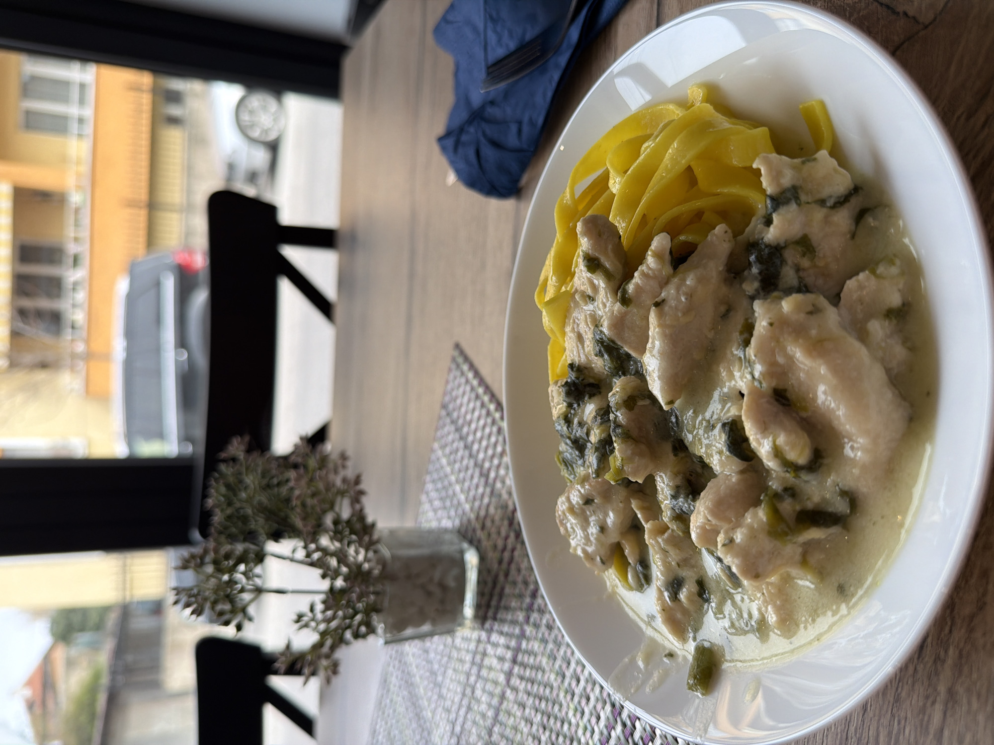 A partially rotated overhead shot reveals a dining table scene, likely inside a restaurant or cafe in Panagyurishte, Bulgaria. A white plate, positioned on the right, holds yellow tagliatelle pasta topped with a creamy, possibly chicken-based, sauce that contains greens like spinach. To the left of the plate, a purple and white patterned placemat sits beneath a small glass vase containing a sprig of dried flowers. A dark blue folded napkin and a fork are visible on the wooden table. The background is a window showing buildings across the street and a dark van. A portion of a black chair is visible between the vase and window. The location is presumed to be in Panagyurishte based on the information provided.