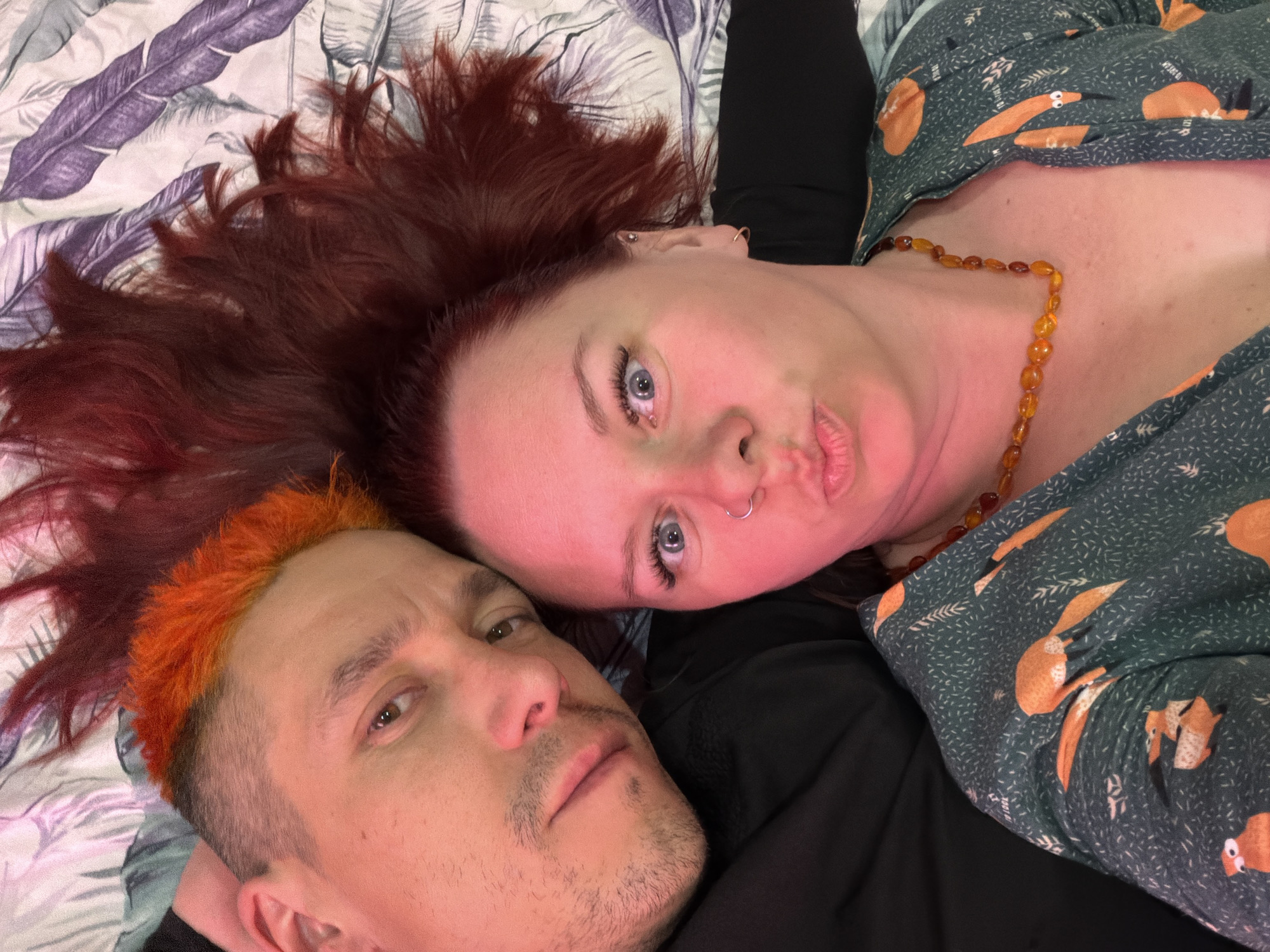 Two people are lying down on a patterned surface, possibly a bed. A woman with auburn hair is positioned at the top of the frame, looking directly at the camera with her lips pursed. She is wearing a dark green shirt with an orange fox pattern and an amber necklace. A man with orange hair is situated on the bottom left, also looking toward the camera with a more serious expression. They appear to be close to each other. The background is a light surface with a purple leaf pattern. It is impossible to determine from the image if they are in the city of Chaikovsky, Russia.