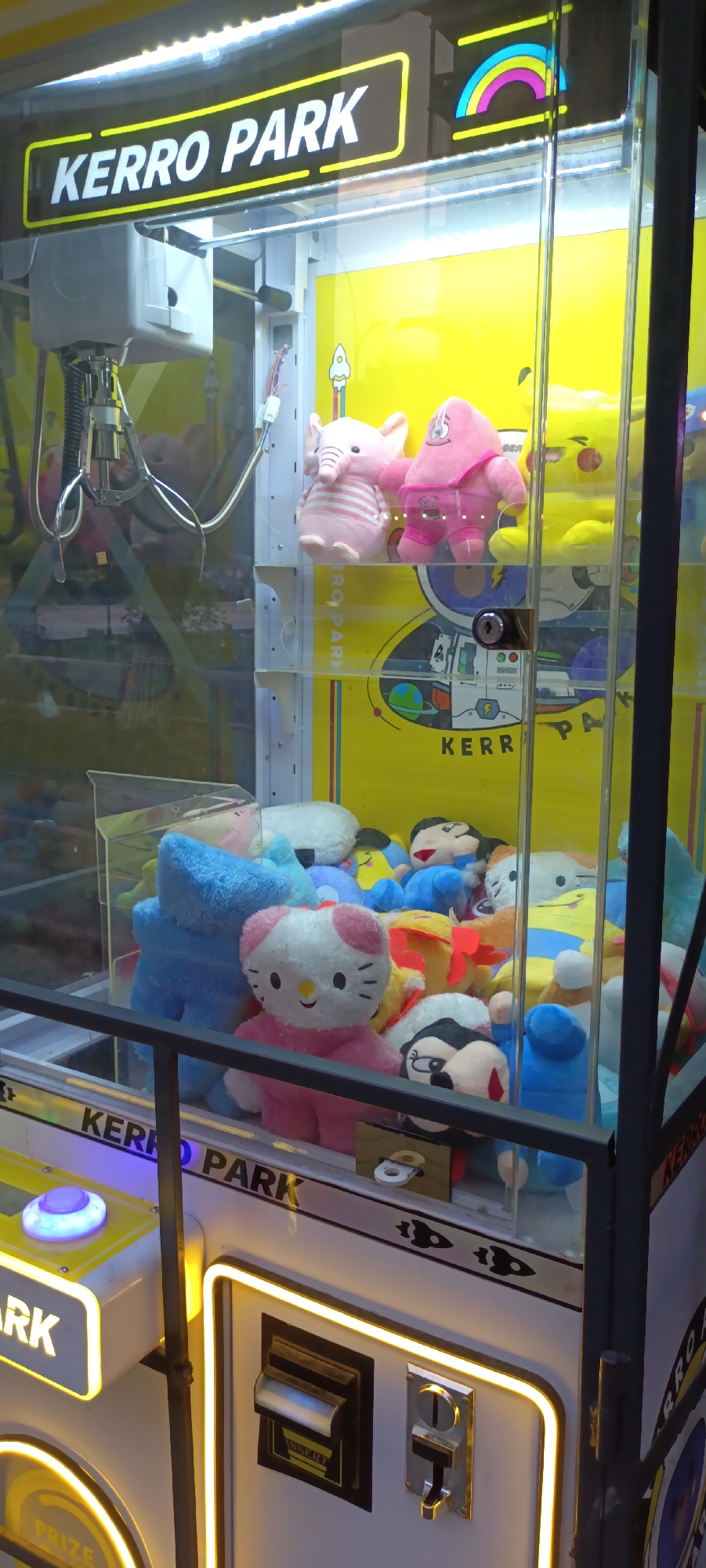 The image shows the interior of a claw machine, branded "KERRO PARK," filled with various plush toys. Prominently featured is a pink Hello Kitty plush. Other plush toys include pink elephants, a pink character resembling Patrick Star, and various cartoon characters. The machine has a control panel with a blue glowing button and coin slots. The background is yellow with graphics, including images of planets and astronauts. The claw mechanism is visible at the top of the machine. The lighting is bright, illuminating the toys within the glass enclosure. The overall scene depicts a typical arcade or entertainment center setting. The location is identified as Fergana City, Uzbekistan, based on the provided context.