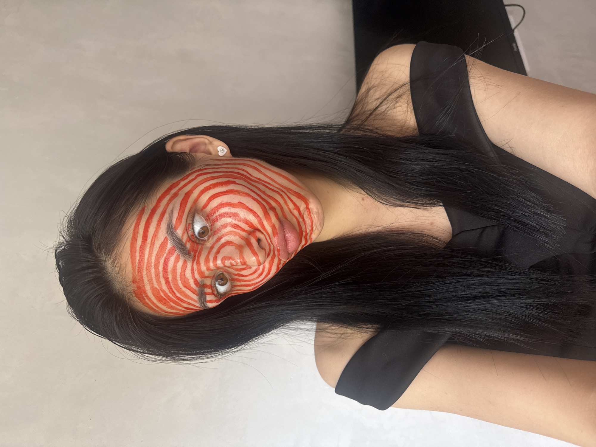 A young adult female with long, straight black hair is depicted in an angled portrait, positioned horizontally against a plain, light-colored background, suggesting an indoor setting. Her face is covered in a striking pattern of intricate, swirling red lines that form concentric circles and curves across her forehead, cheeks, nose, and chin. These bold red lines create a hypnotic and artistic effect, leaving her brown eyes, eyebrows, and lips largely unpainted, though some lines extend close to these features. Her eyes are wide open, gazing upward and slightly to the right, and her lips are slightly parted. She wears a small, white, heart-shaped ear stud in her left earlobe, which has the text "ME" inscribed on it. Her attire includes a dark, possibly black, top or dress with a broad strap, visible around her shoulder and chest. The overall impression is a dramatic and intense artistic presentation, likely for a photoshoot showcasing the elaborate face paint. The scene is set in Votkinsk, Russia.
