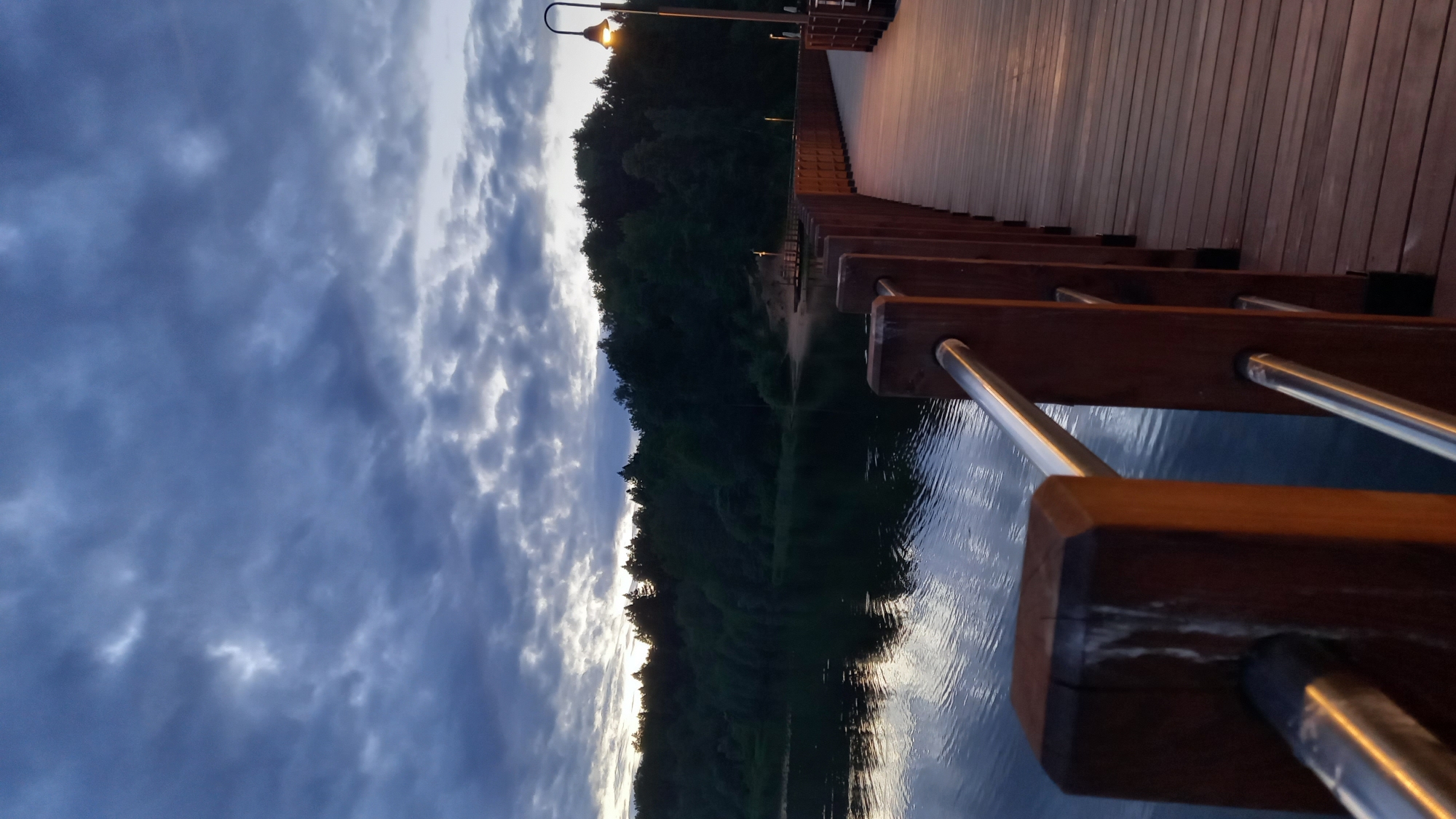 The image captures a waterside scene in Krasnogorsk, Russia, featuring a wooden pier or walkway extending over a body of water. The structure has railings with wooden posts and metal bars. A light post stands at the beginning of the pier. The water reflects the sky, which is filled with dark, cloudy formations at dusk or dawn. Across the water, a dense forest line is visible.