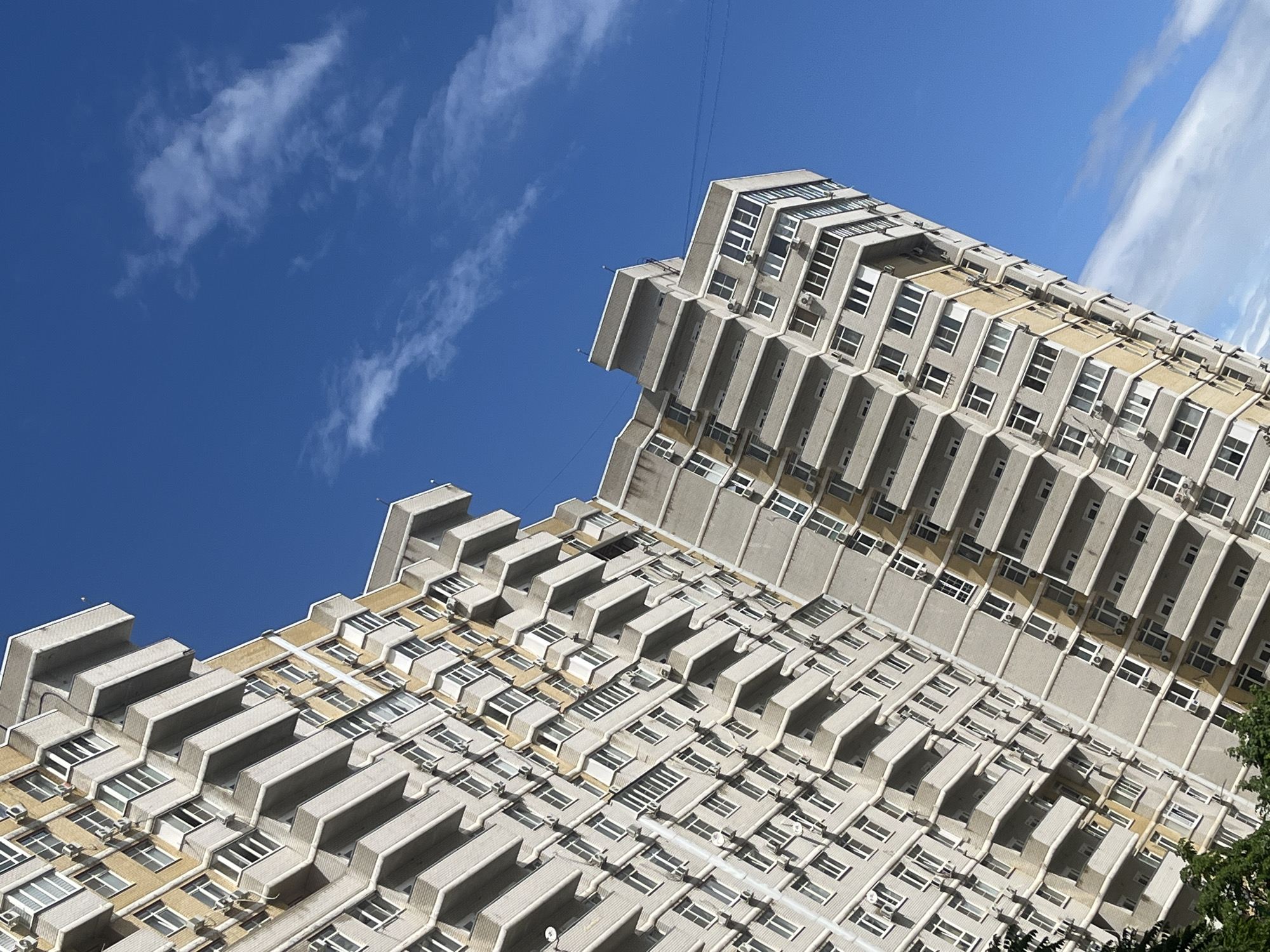 The image shows a low-angle shot of a tall, multi-story building under a partly cloudy blue sky. The building has a distinctive zigzag pattern, with each level jutting out slightly from the one below. The structure appears to be made of concrete or a similar material, with rows of windows and air conditioning units visible on each floor. The building is located in Krasnodar, Russia.