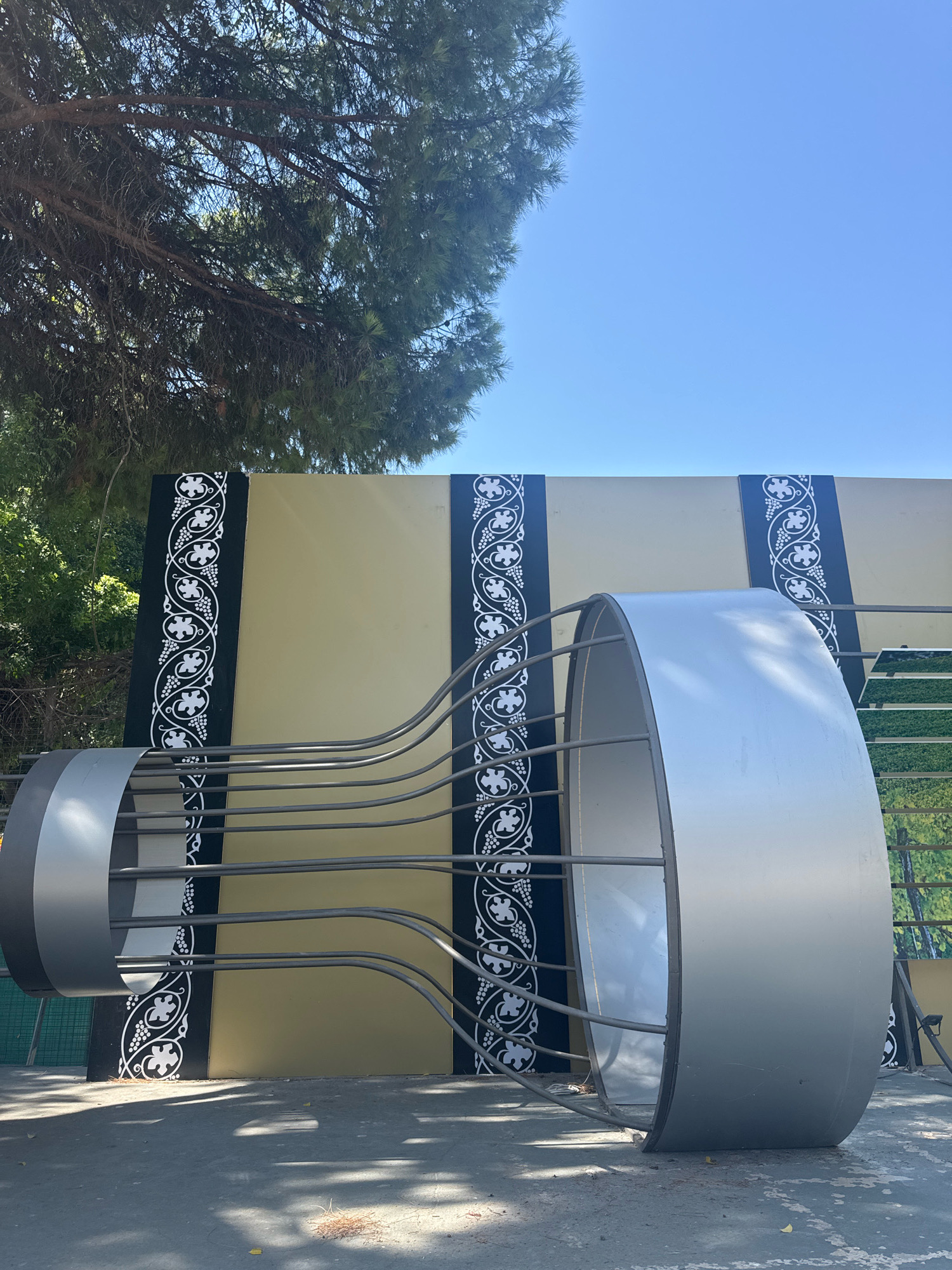 The image depicts an outdoor art installation in Limassol, Cyprus. The installation features a large, abstract sculpture made of metallic elements. The sculpture is cylindrical and appears to be composed of two large rings connected by numerous thin, curved metal rods. The background consists of a large, flat panel with a mustard-yellow surface, bordered by decorative black panels with a white, vine-like pattern. The scene is set on a paved surface, with shadows cast by sunlight and overhead foliage, suggesting a daytime setting. A portion of a green tree with pine needles is visible in the upper left corner of the frame, and a clear blue sky dominates the upper portion of the image. The overall impression is that of a modern artistic display in a public space, likely outdoors.