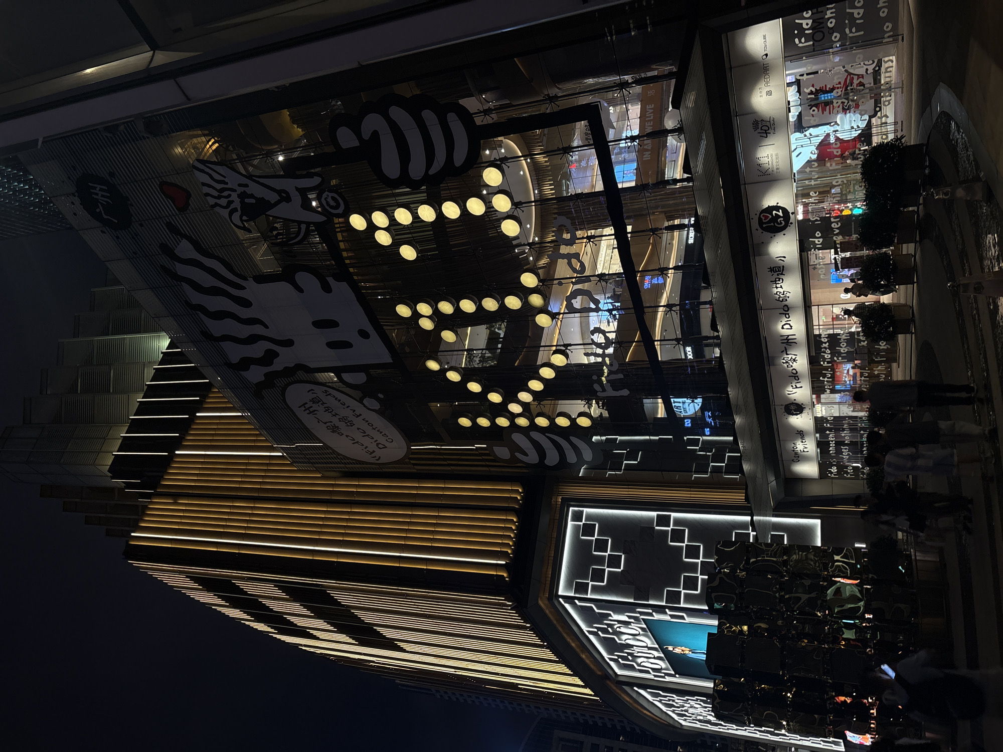 This is a nighttime, exterior shot of a modern shopping complex. The building features a striking architectural design with horizontal lighting accents on some sections, creating a warm glow against the dark sky. The facade incorporates large glass windows, revealing illuminated interiors with displays and what appear to be mannequins or decorative figures.
Prominently displayed on the glass are various logos and text, including "K11", "GZ", and "BREDART". There is also Chinese text such as "艺文化地标" (Art and Culture Landmark) and "主办方" (Organizer). English text like "Canton Friends" and phrases starting with "Fido" are visible, suggesting promotional material or brand messaging. A large black and white graphic, possibly of a dragon or stylized creature, adorns a section of the building.
Several people are visible in the foreground and on the steps leading up to the complex, appearing to be shoppers or passersby. They are mostly silhouetted against the lights, with their activities not clearly discernible. The scene conveys a sense of urban activity and modern commerce at night, with the building itself being a focal point due to its distinctive lighting and design elements. The reflection on the glass surfaces adds another layer of visual complexity to the scene.